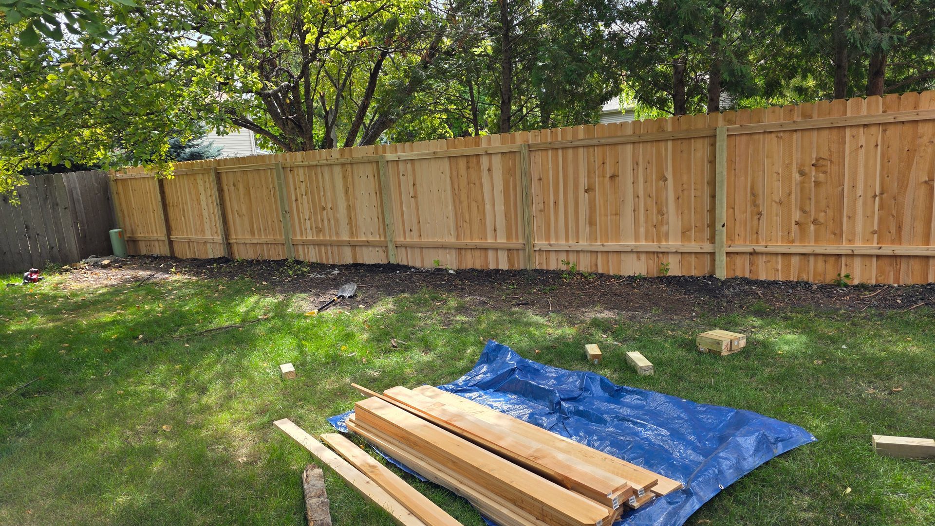 Wooden fence in a backyard, construction materials on a blue tarp, grass, trees.