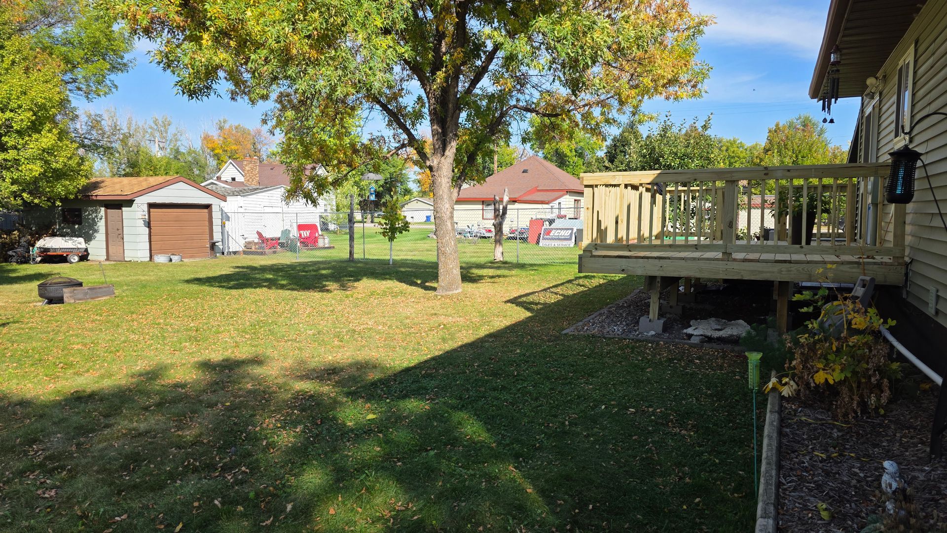 Backyard with a wooden deck, tree, garage, and houses under a clear blue sky.