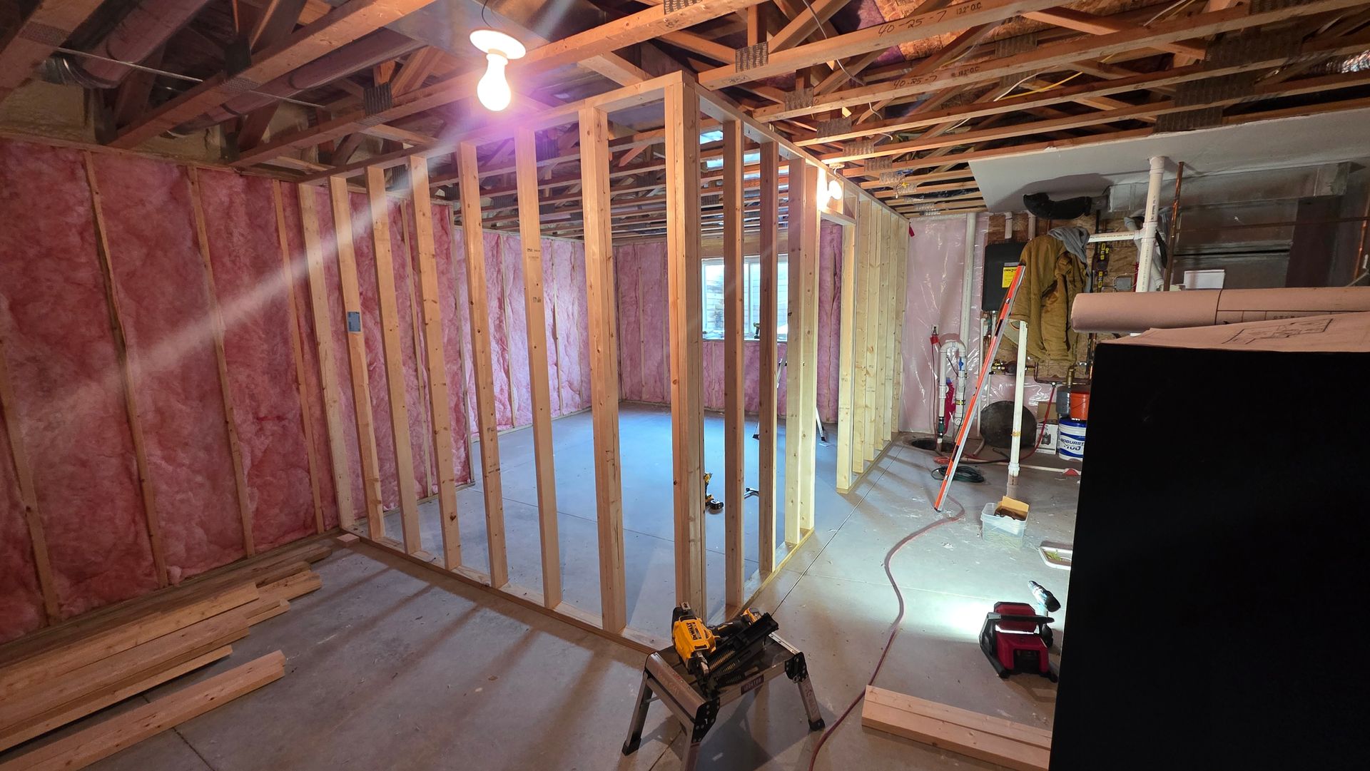 Basement renovation in progress. Wood framing, pink insulation, tools, and exposed ceiling beams.