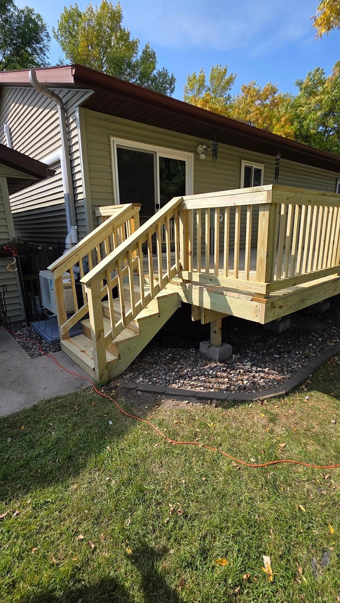 Wooden deck with stairs attached to a green house, surrounded by grass and trees.