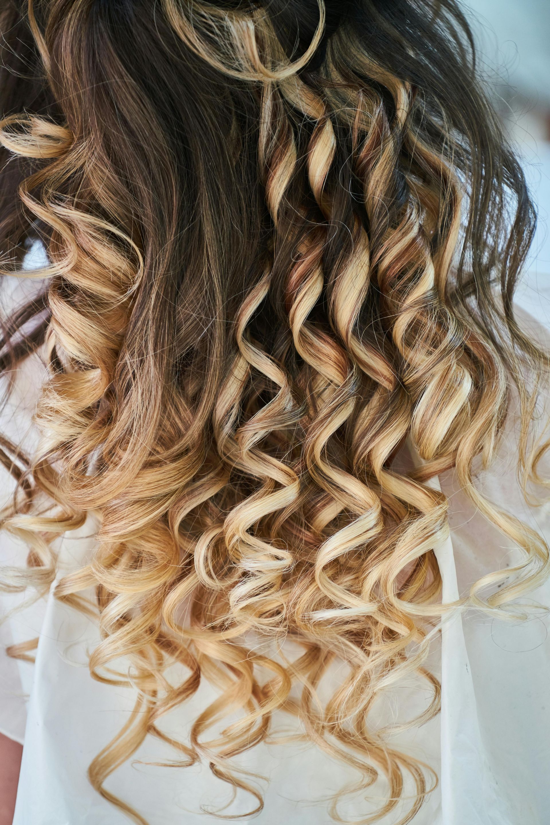 Shoulder-length hair with brown roots and golden blonde highlights, styled into defined, spiral curls.