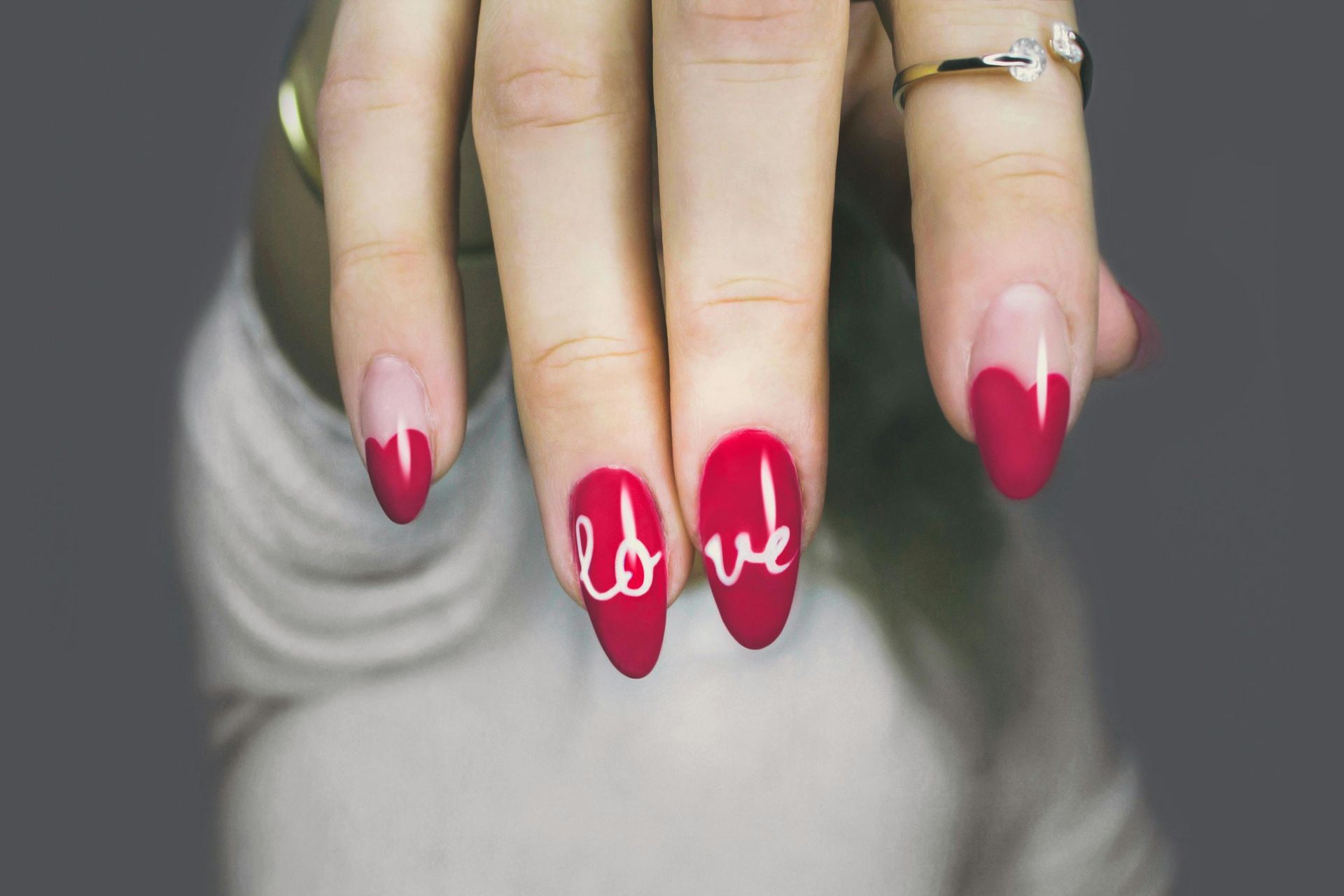 A close-up of a hand with red, heart-shaped French manicure nails featuring white cursive letters spelling out