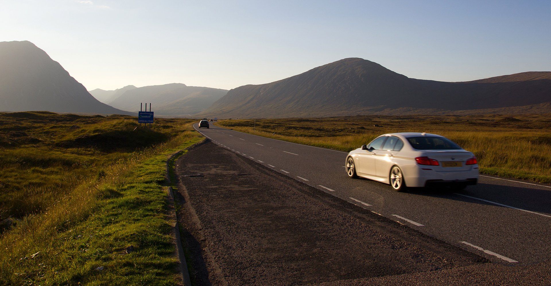 A white car is driving down a road with mountains in the background.