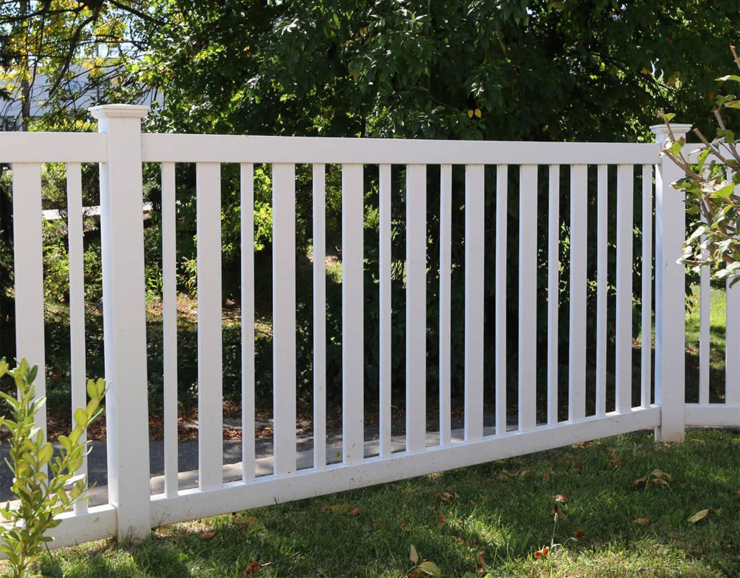 White vinyl fence with vertical slats in a grassy yard, backed by trees and shrubs