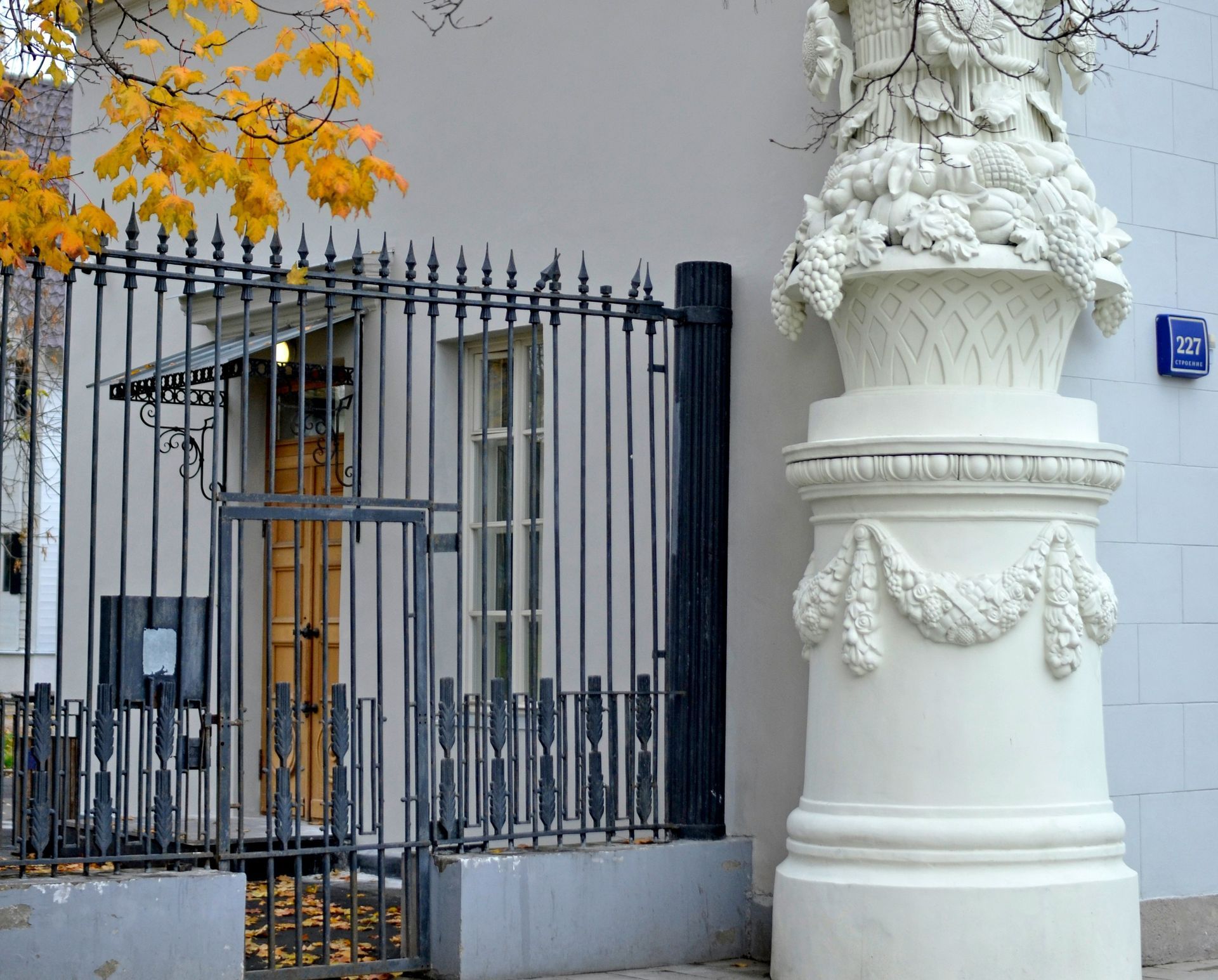 Exterior of a building with a white decorative column, metal fence, and autumn leaves.