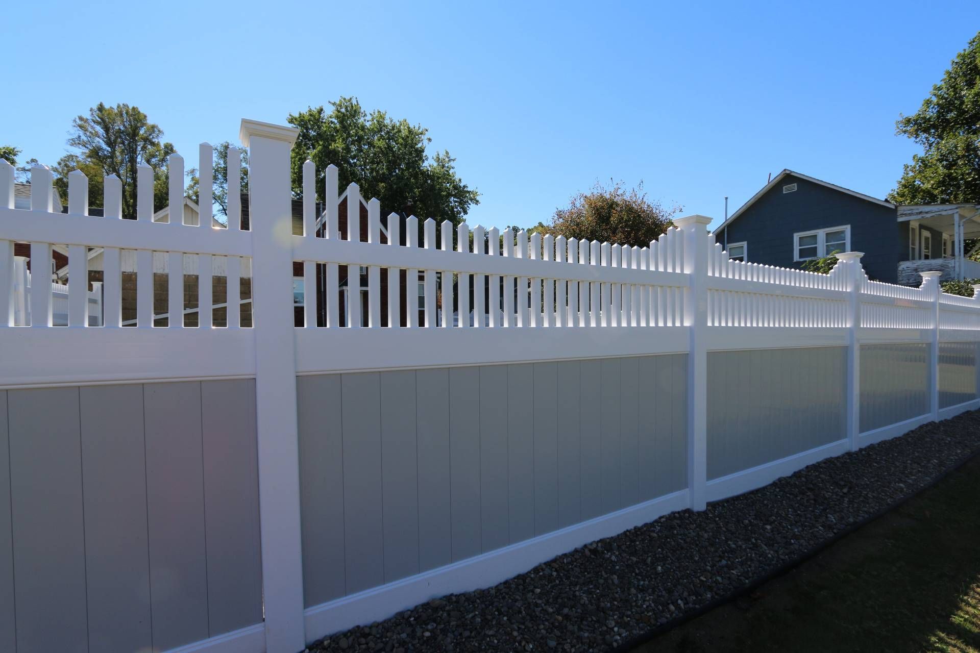 White vinyl fence along a sloped yard under a blue sky, with houses and trees in the background