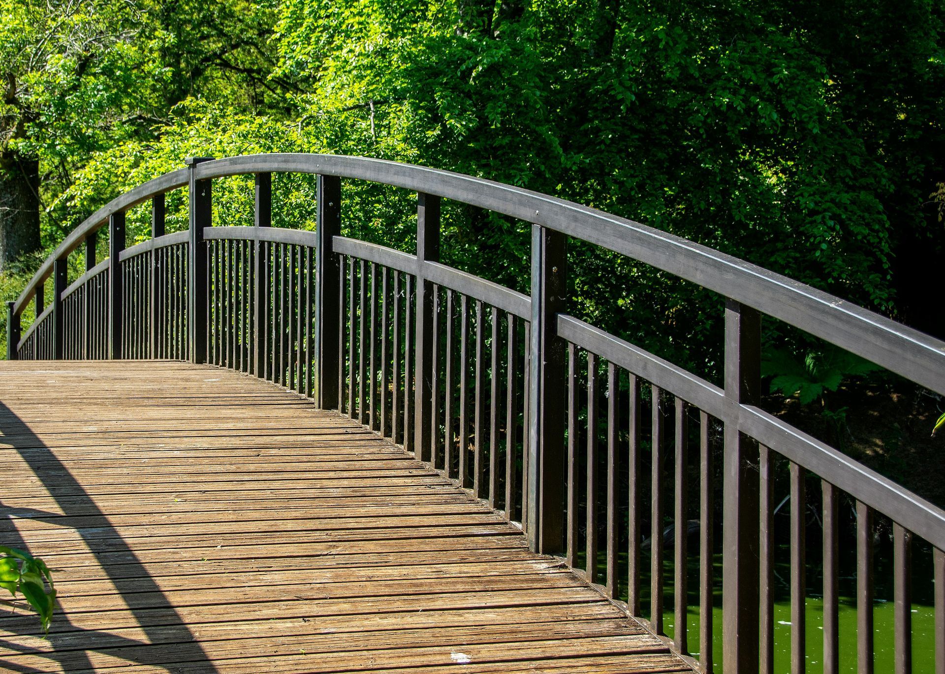 Wooden arched bridge with black railings over water, lush green trees in the background.