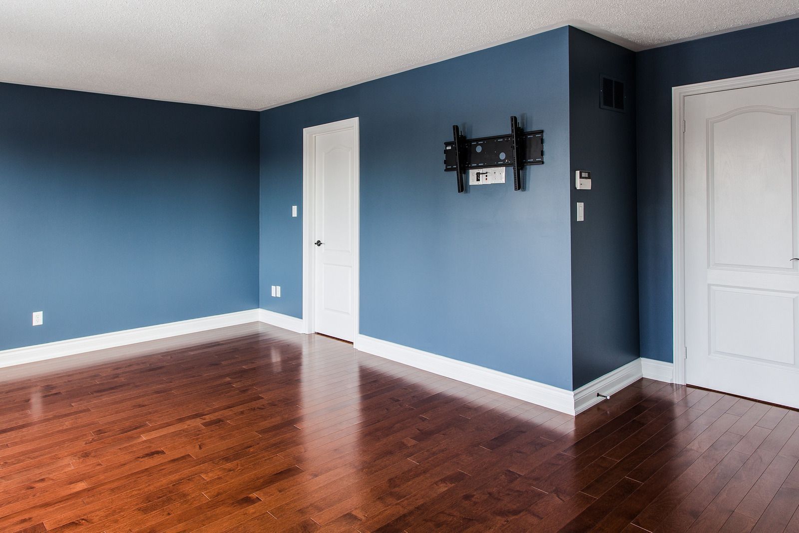 A room with blue walls, dark wood floors, white trim, two white doors, and a TV wall mount installed on the blue wall.