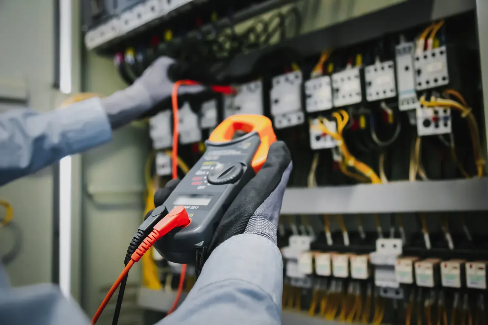 A person in work gloves uses a digital clamp meter to test electrical components inside an industrial control panel.