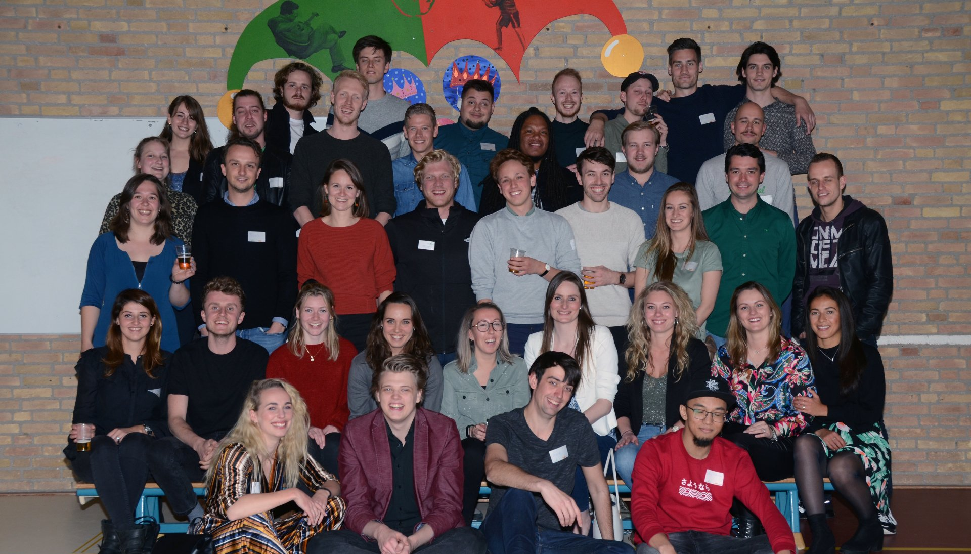 Groepsfoto van diverse mensen in een kamer, lachend, sommigen met naambordjes. Kleurrijke achtergrond.