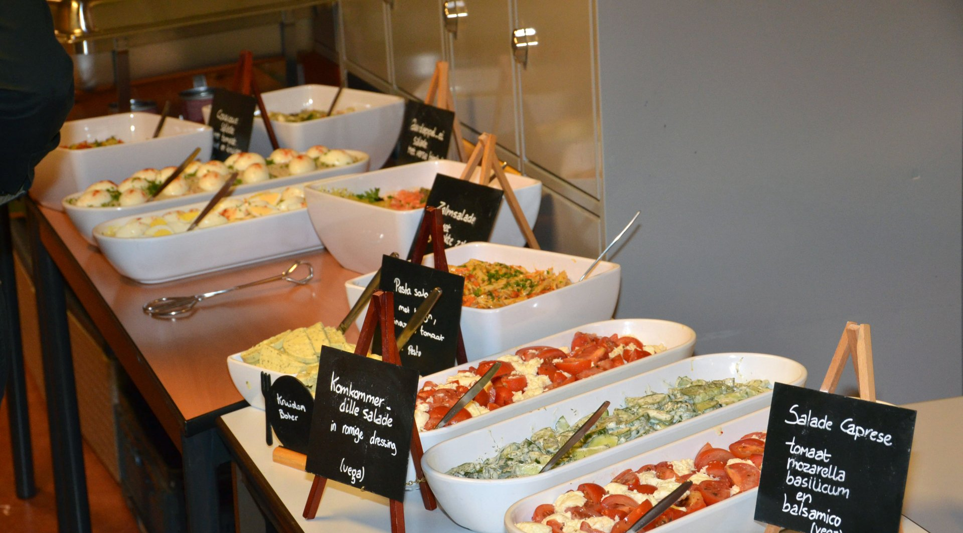 Buffet met diverse salades in witte serveerschalen, aangegeven met kleine zwarte bordjes.