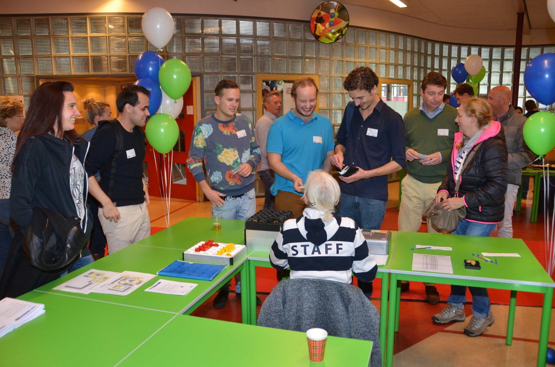 Mensen aan een tafel met een personeelslid in een gestreept shirt. Ballonnen en felgroene tafels.