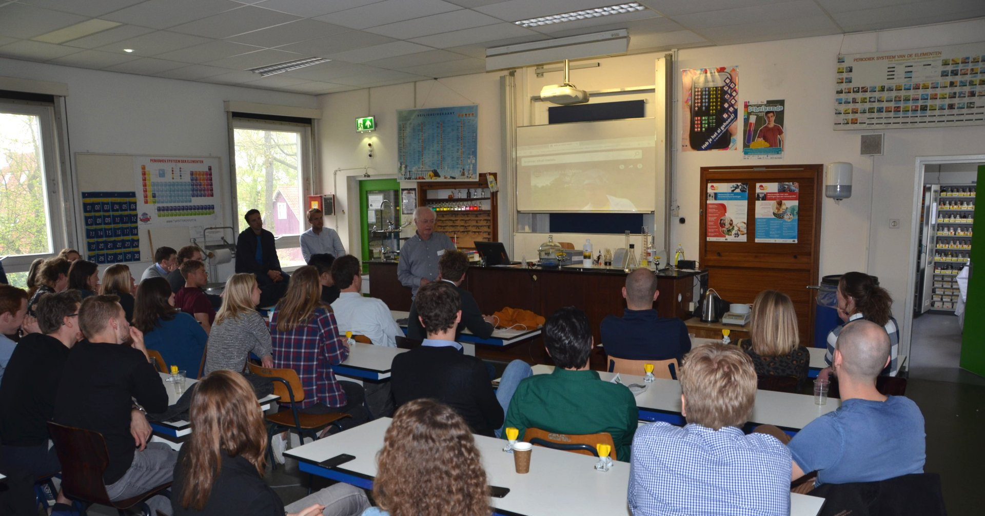 Een groep mensen in een kamer luistert naar een man die een lezing geeft, mogelijk in een klaslokaal.