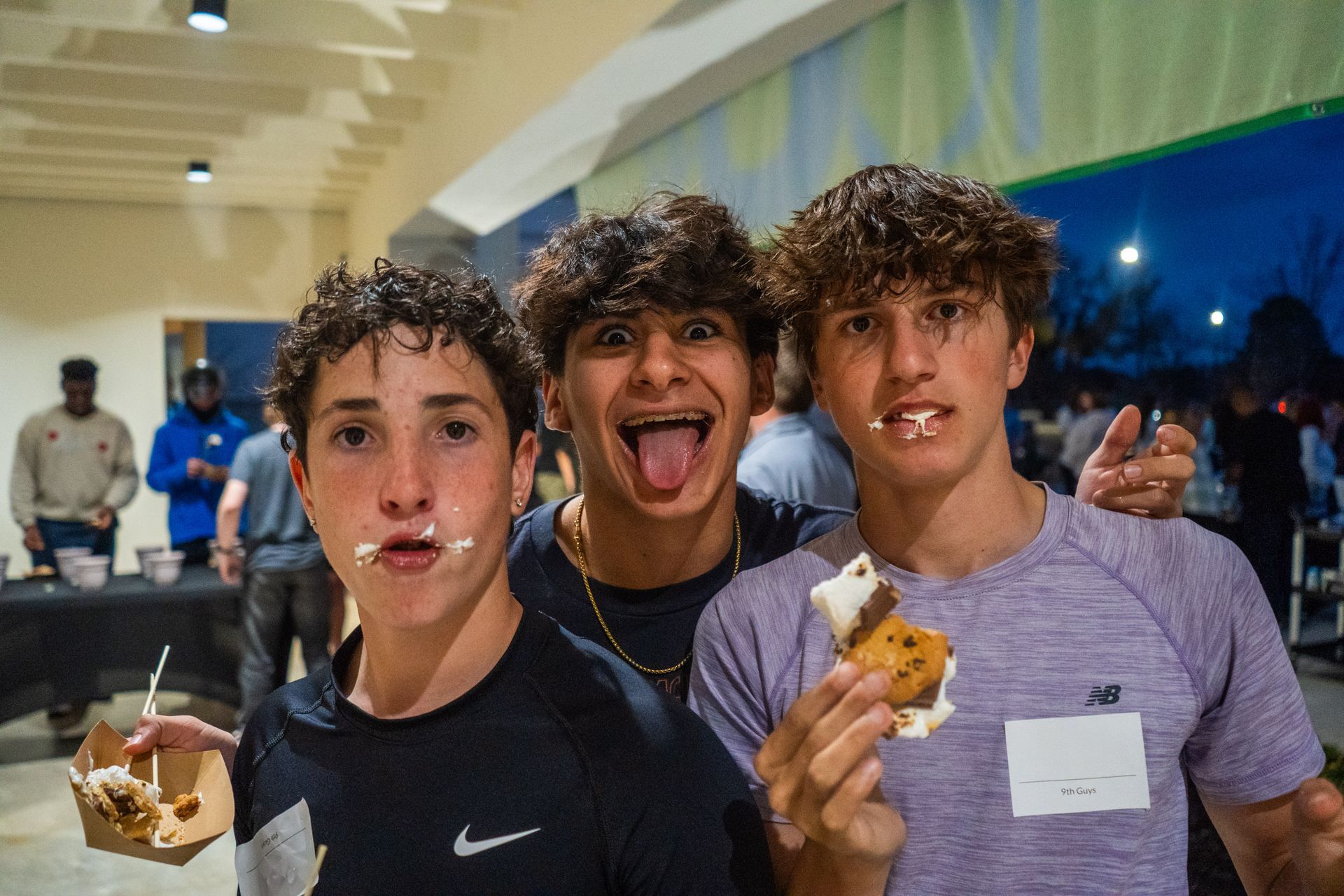 Three young men with messy faces, holding desserts, smiling at camera. Indoor event.