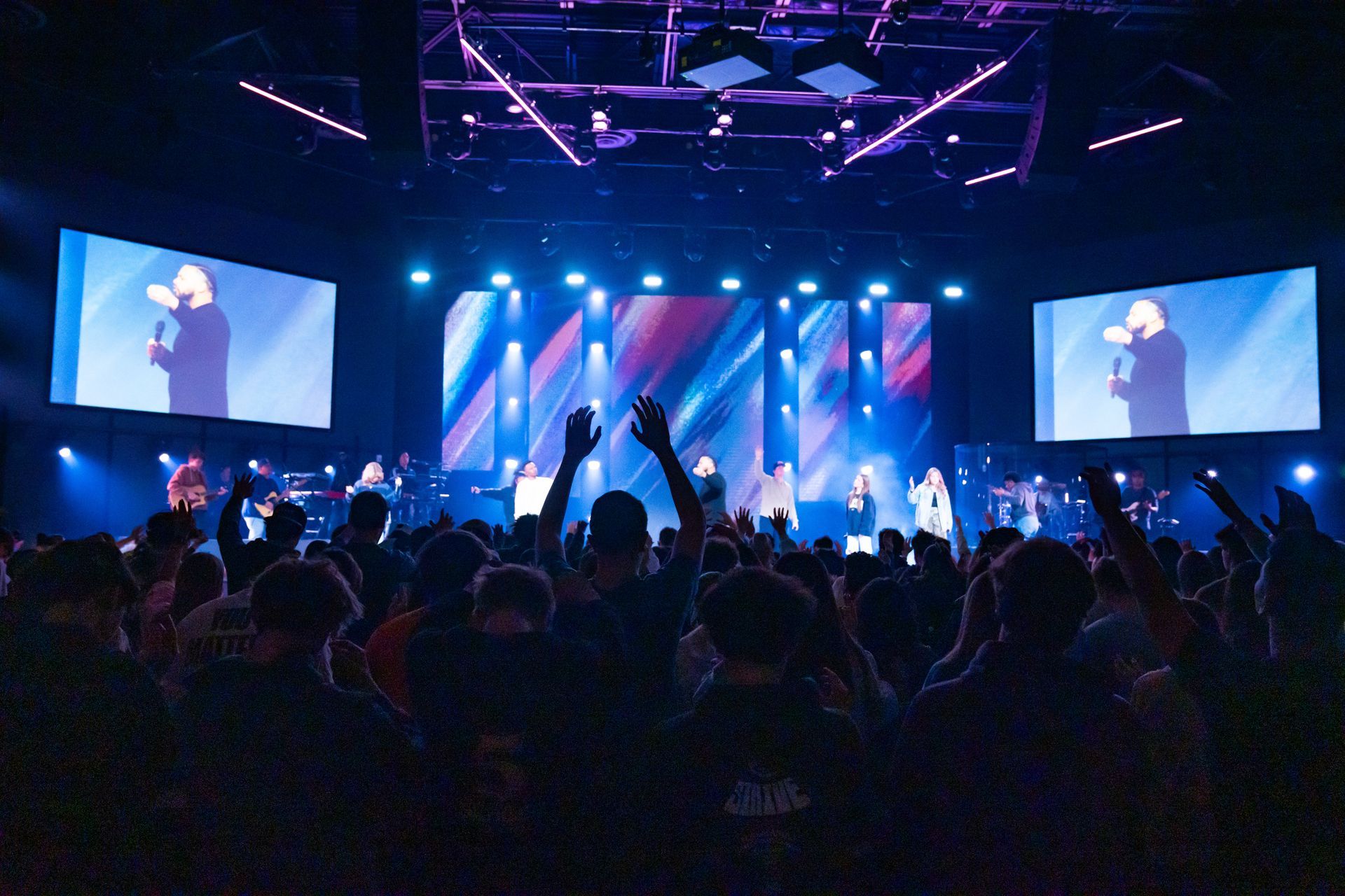 Audience with hands raised at a brightly lit concert; screens show a man speaking onstage.