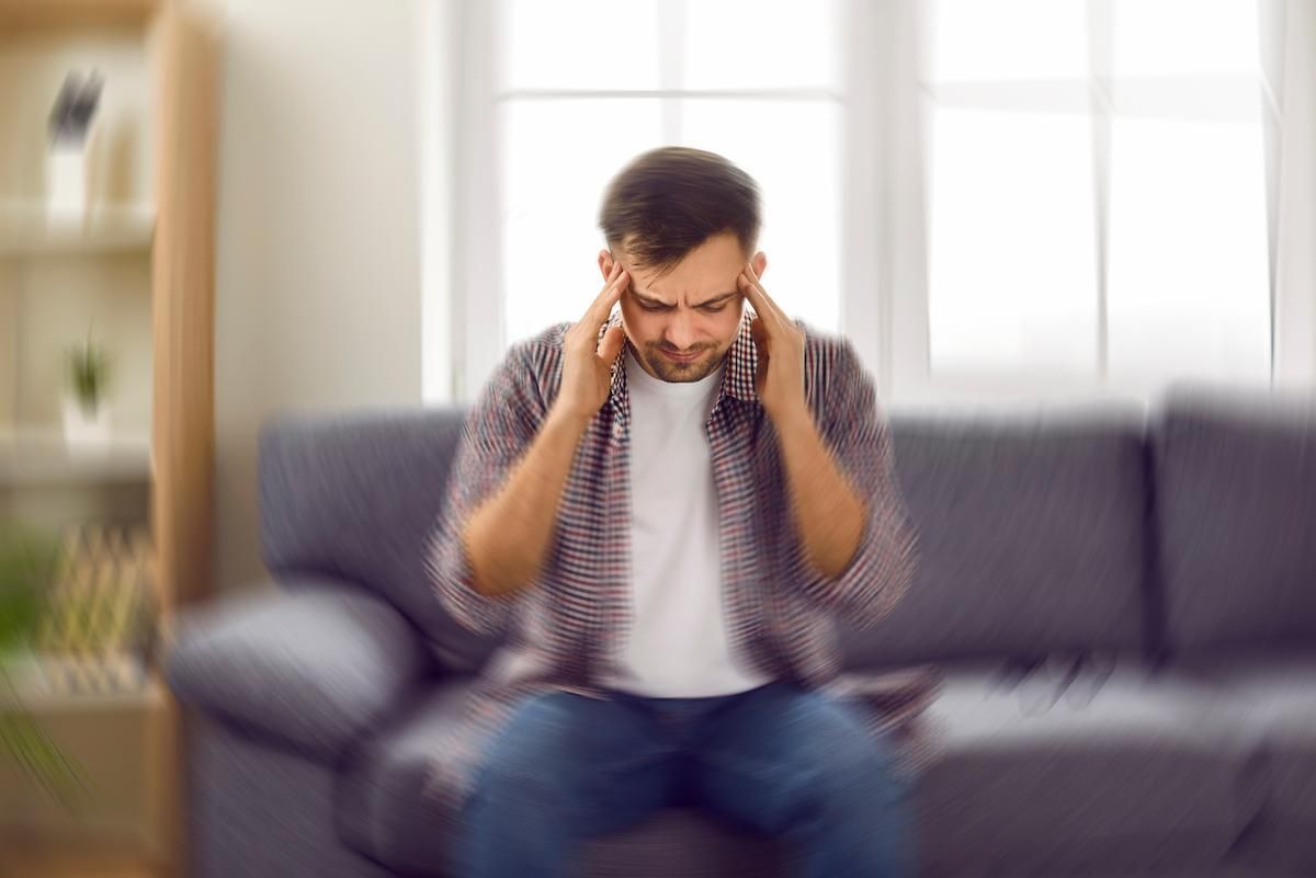 A man sitting on a sofa holds his temples, grimacing from a headache, with a motion blur effect.