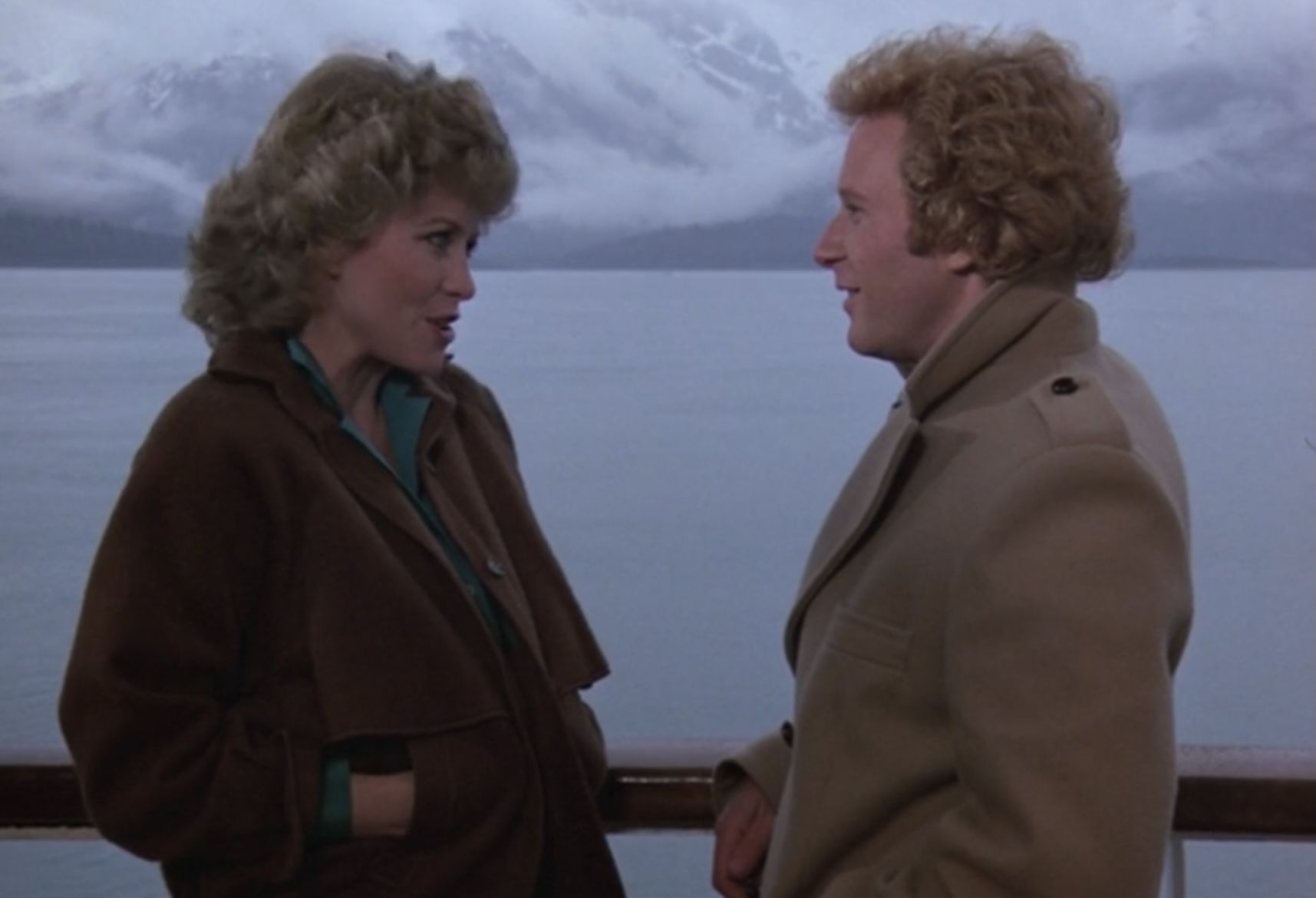 Woman and man conversing outdoors on a boat deck, mountains and water in the background.