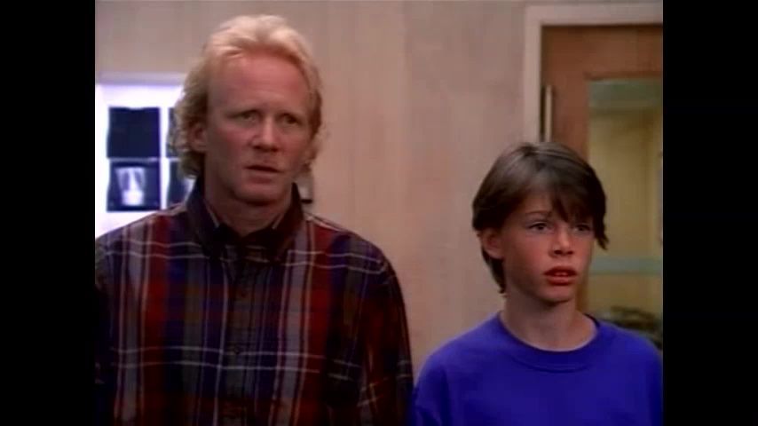 Man and boy with concerned expressions, inside a building. The man wears a plaid shirt, the boy a blue sweater.