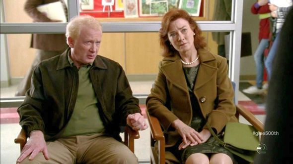 Two people sitting, looking concerned. Man in jacket, green shirt, khakis. Woman in brown coat, green dress.