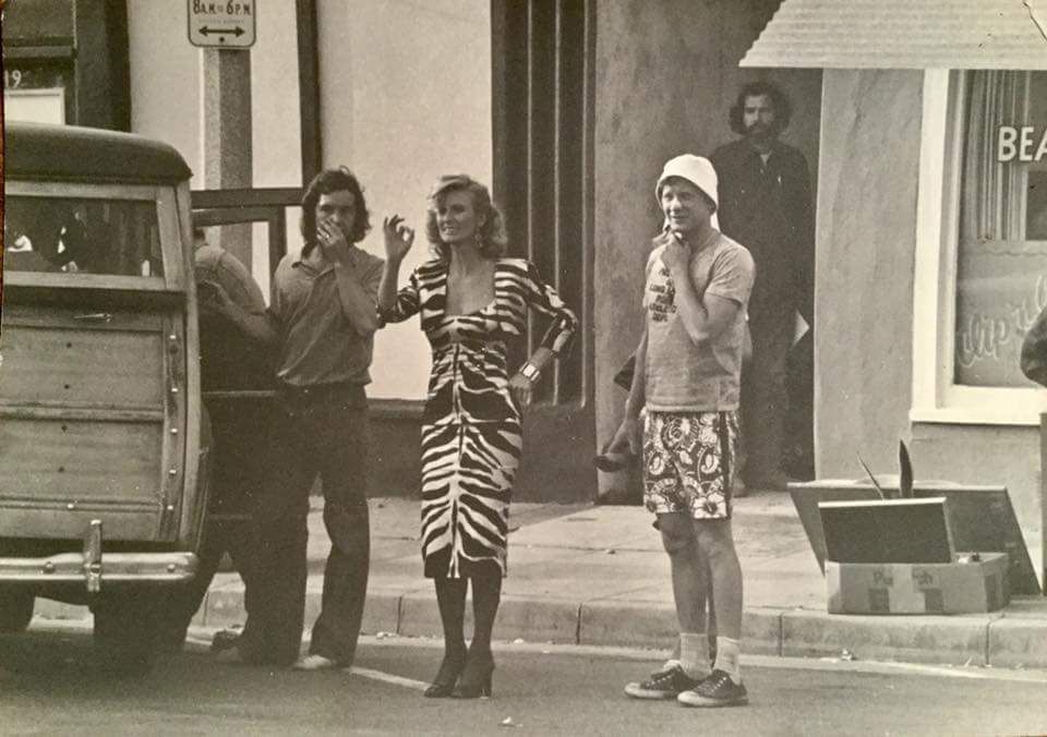 Three people smoking on a street; a wooden-paneled car, building and sign in the background.
