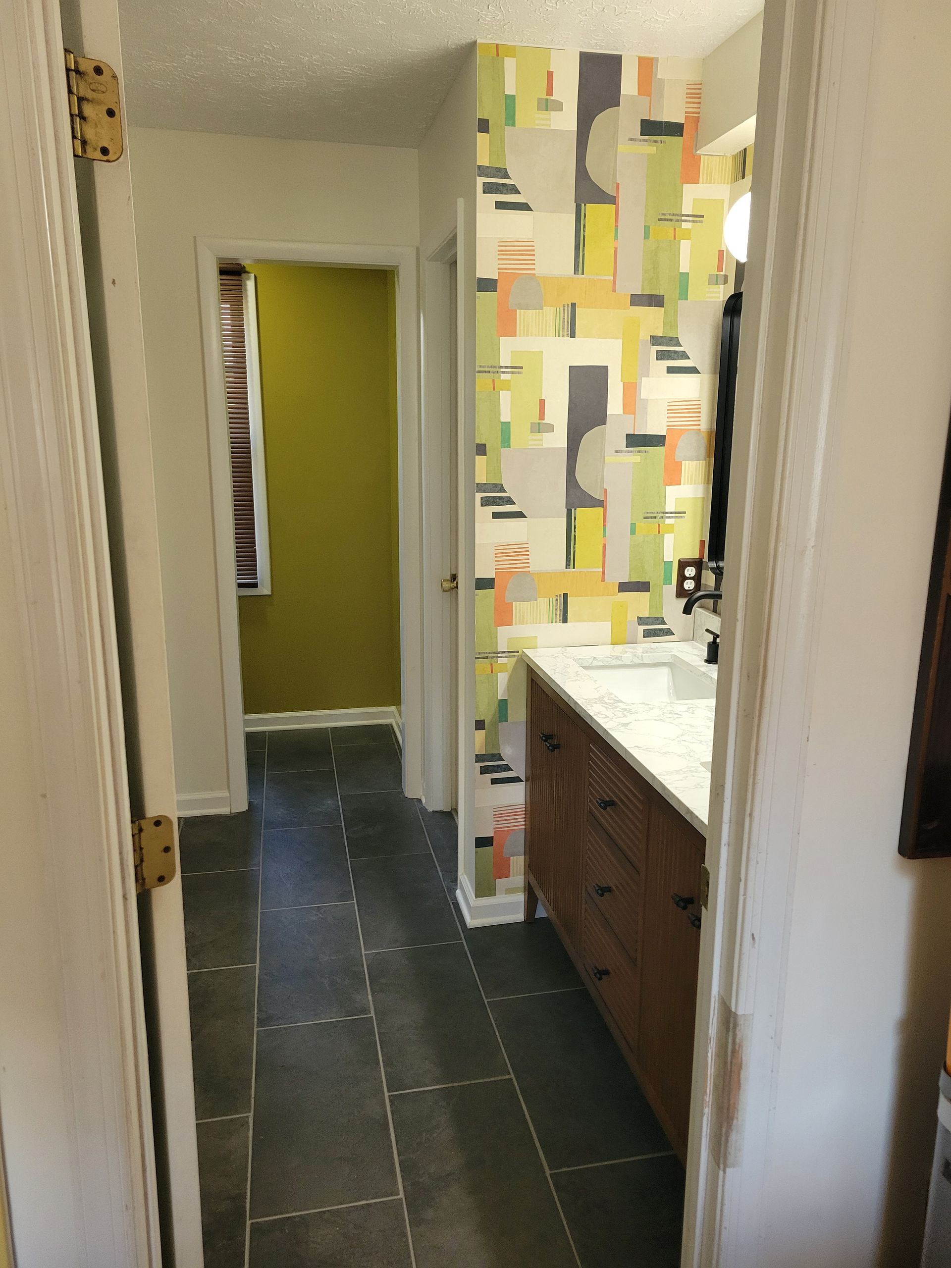 Narrow hallway leading to a bathroom with green walls, dark tile floor, and a vanity with patterned wallpaper.