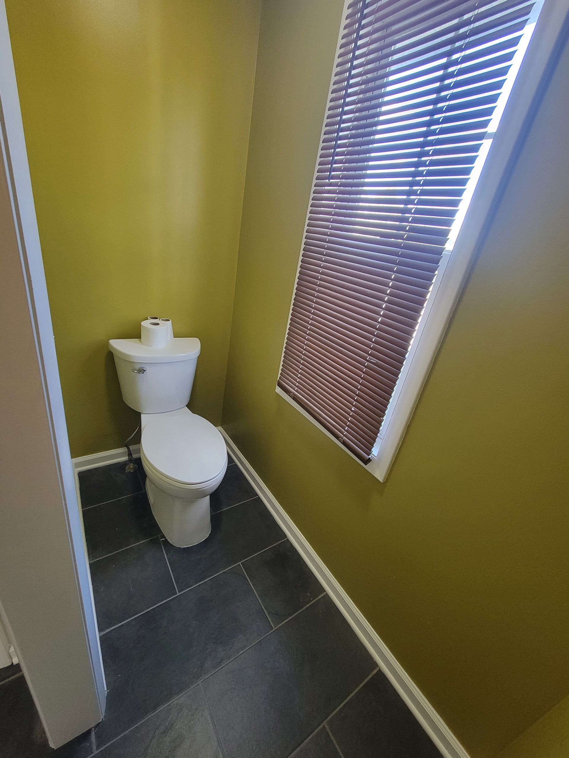 Small bathroom with a toilet, dark tile floor, yellow walls, and a window with blinds.