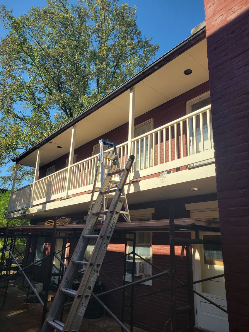 Worker on a ladder repairing a second-story porch with white railings and red brick walls