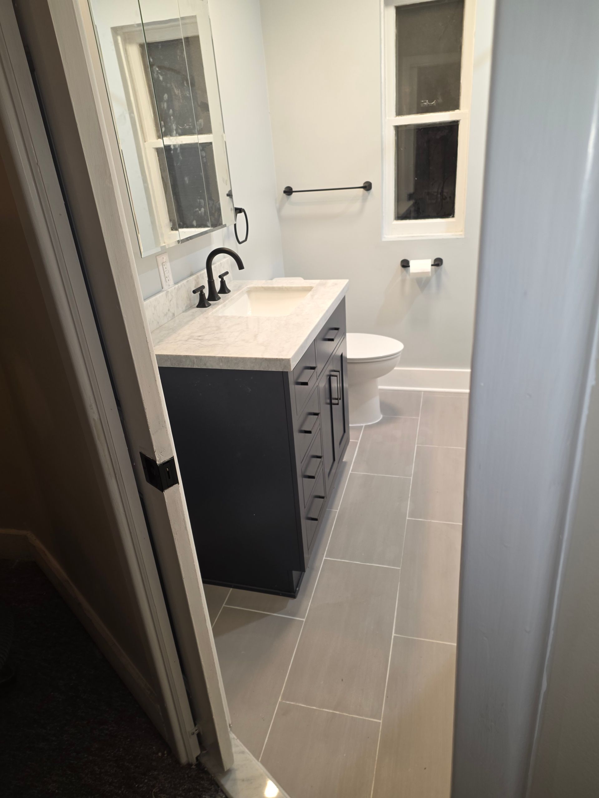 Narrow bathroom with gray tile, vanity and sink, mirror, toilet, and window.