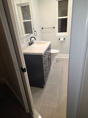Small bathroom with white vanity, black cabinet, toilet, window, and gray tile floor.