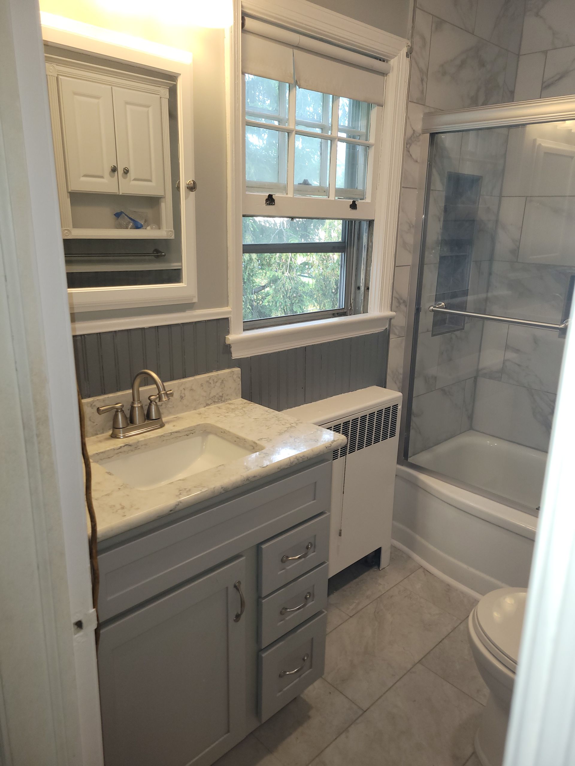 Small bathroom with vanity, mirror, window, and tiled tub shower with glass doors.