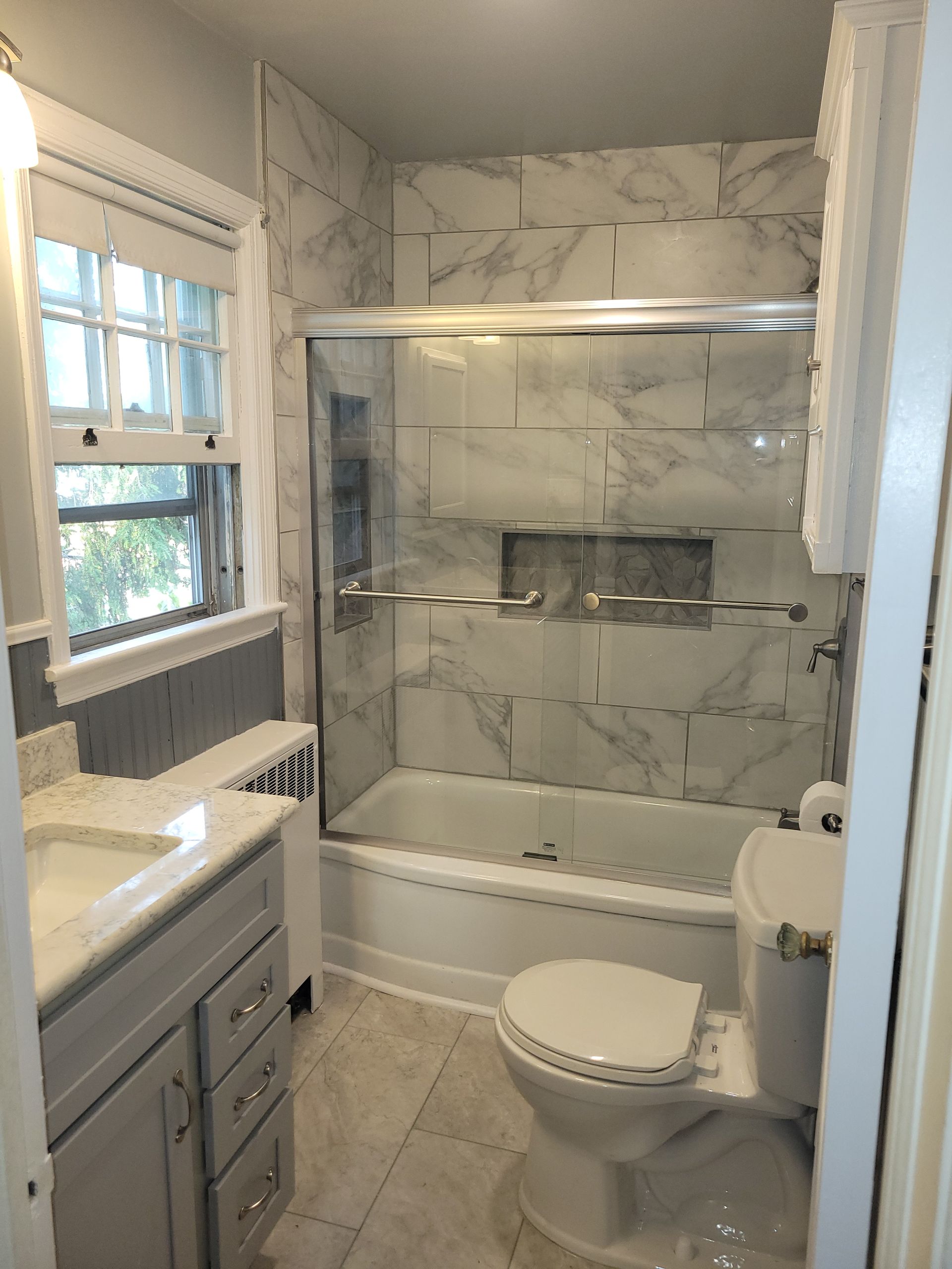 Small bathroom with tub-shower, toilet, vanity, window, and marble-look wall tile.