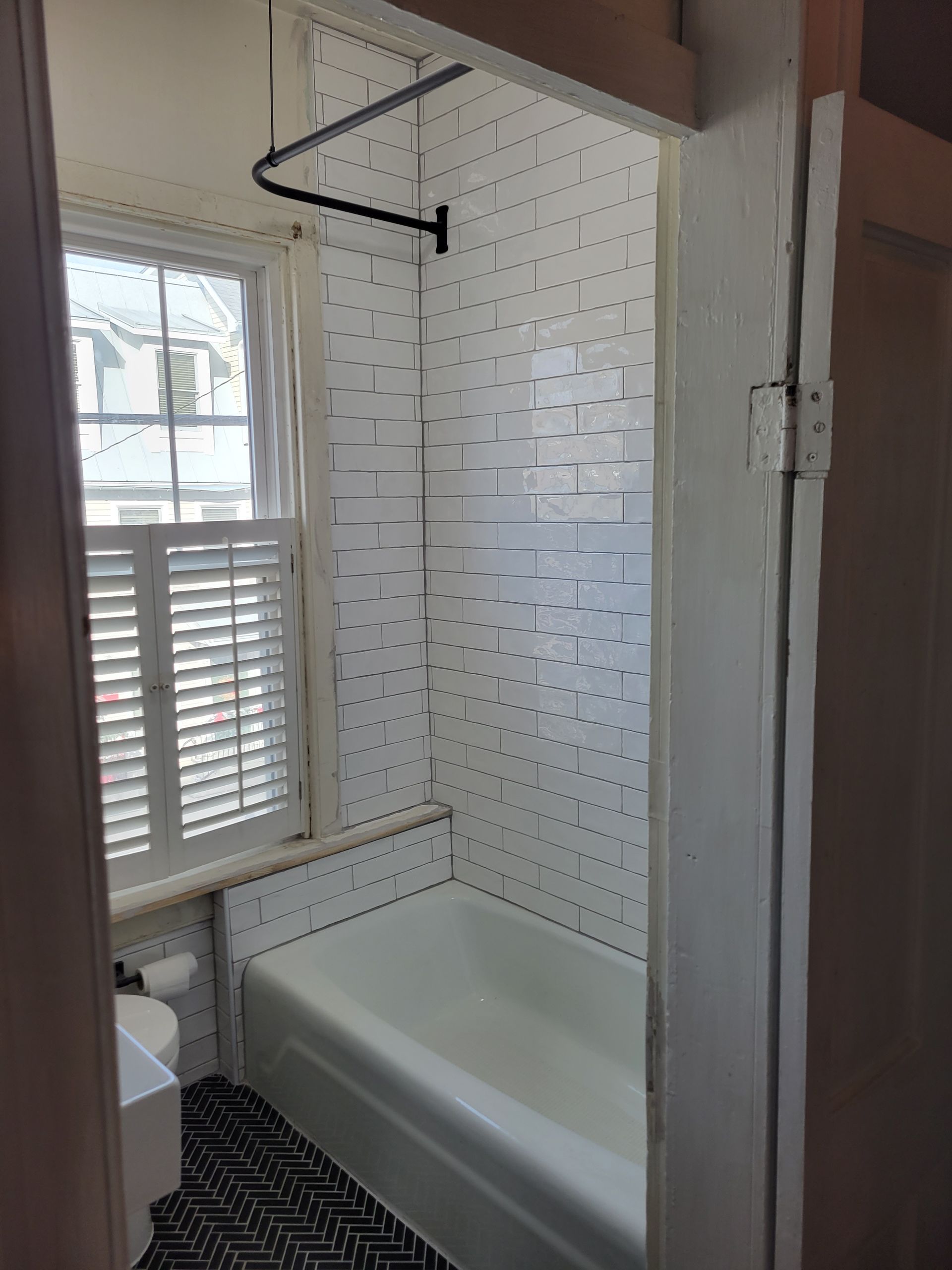Small bathroom with white tiled shower, bathtub, window, and black shower curtain rod