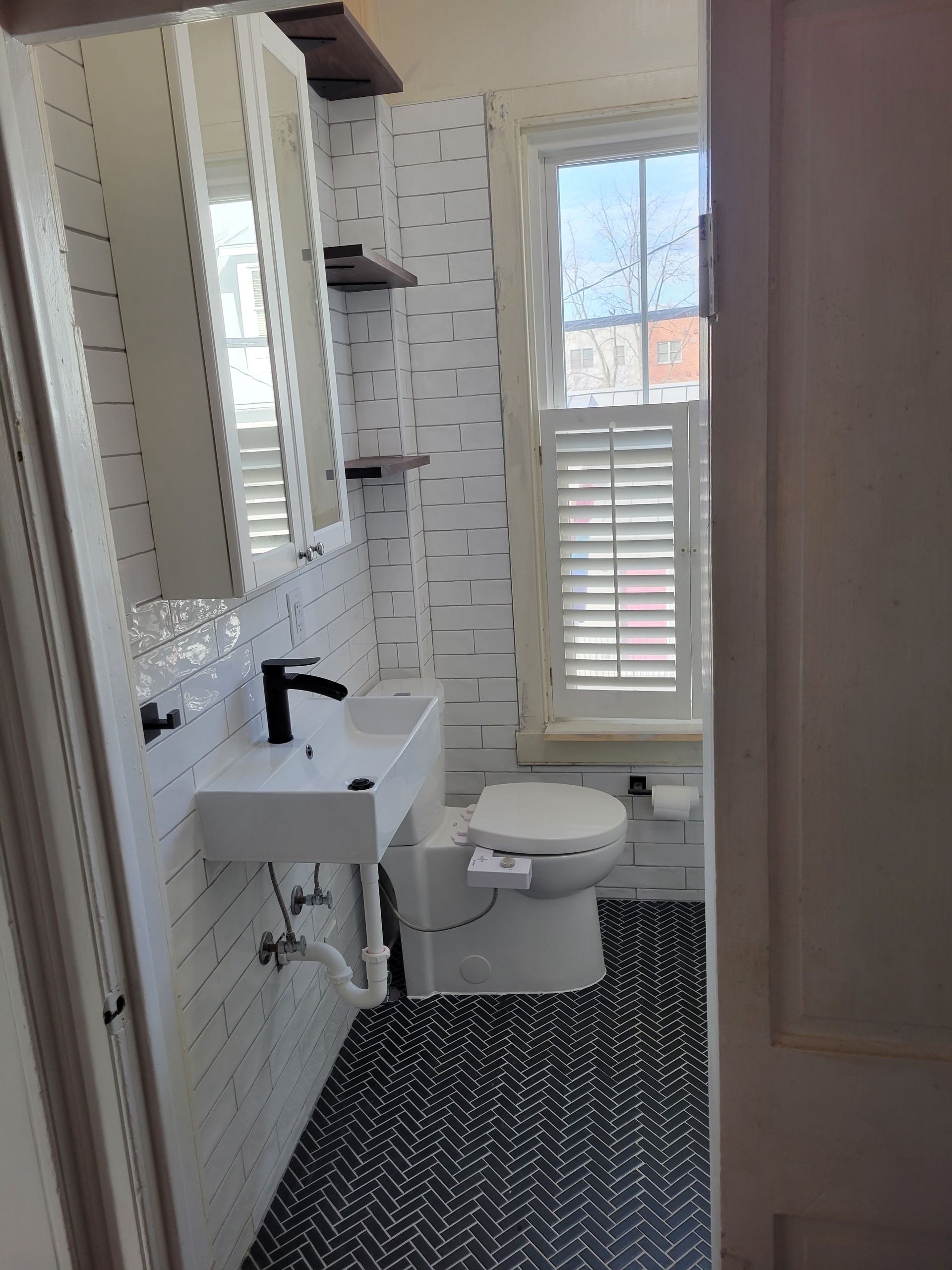 Small white bathroom with pedestal sink, toilet, mirror cabinet, and window with shutter blinds.