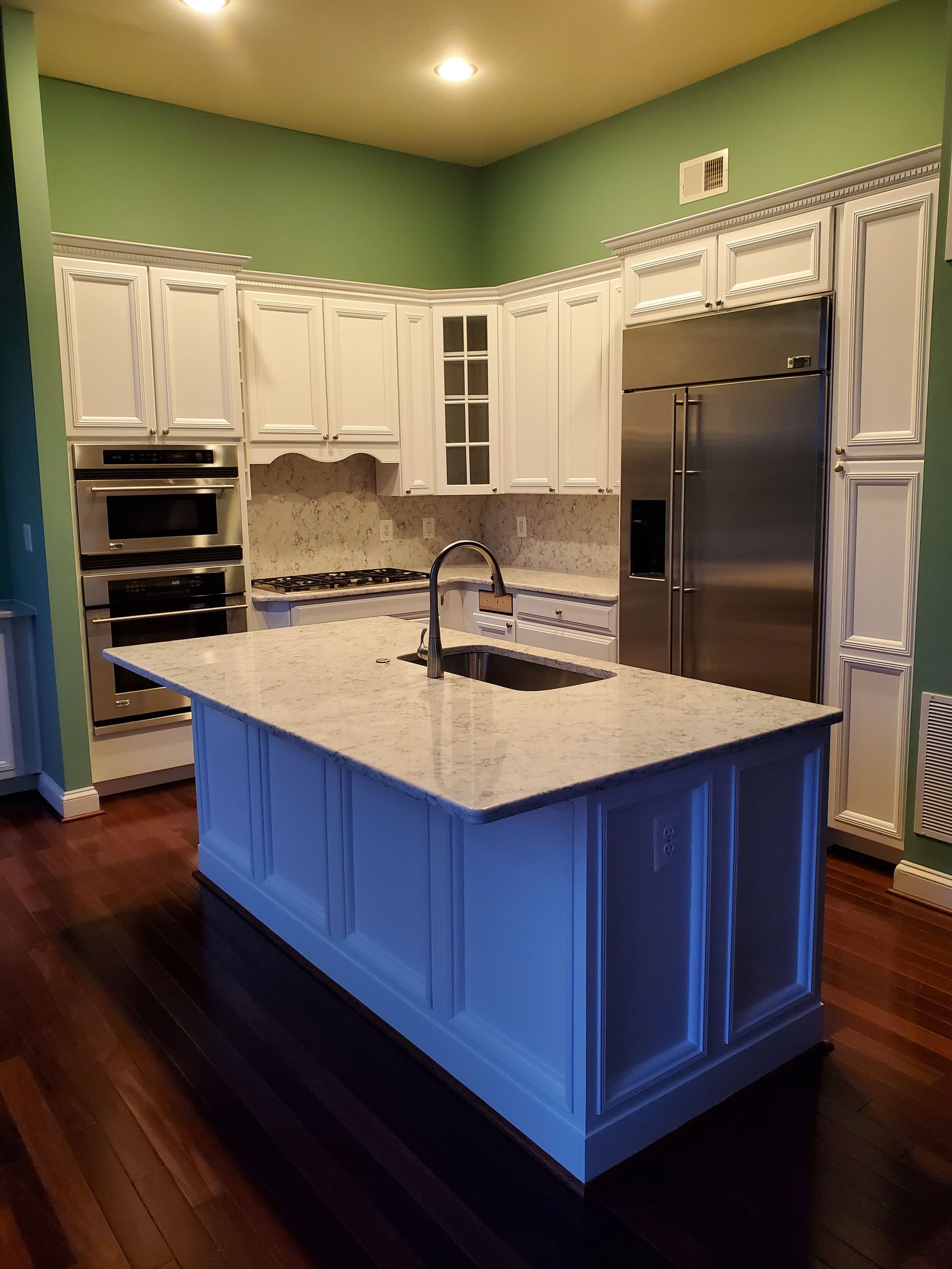 Bright kitchen with white cabinets, green walls, dark wood floors, and a blue island with a sink.