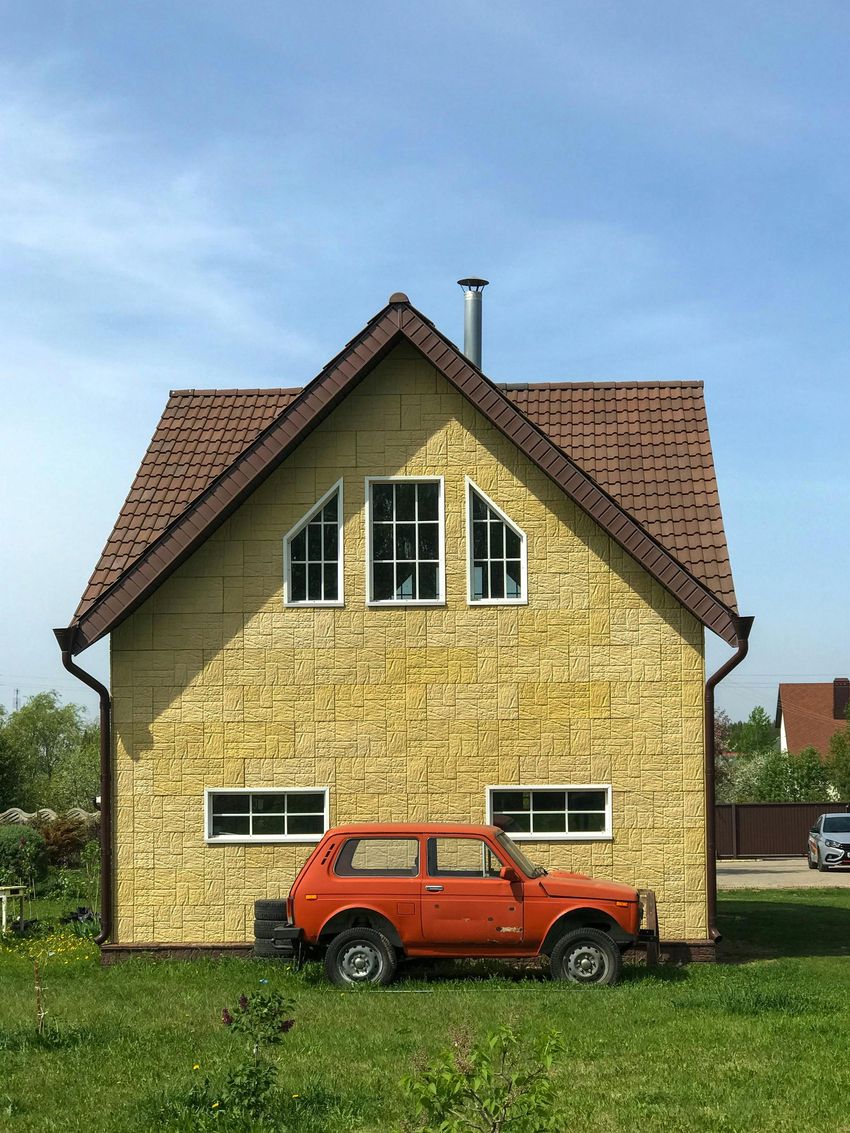 Orange SUV parked in front of a beige house with a steep brown roof and triangular windows.
