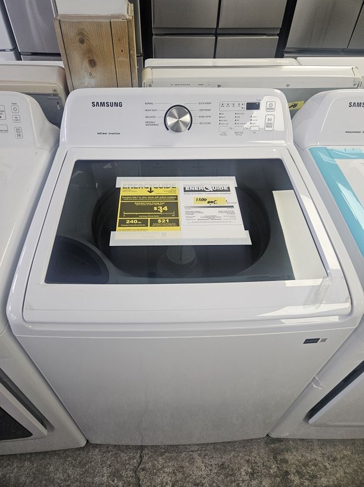 Machine à laver Samsung blanche à chargement par le haut avec couvercle en verre, commandes sur le dessus et étiquette indicateur de consommation d'énergie.