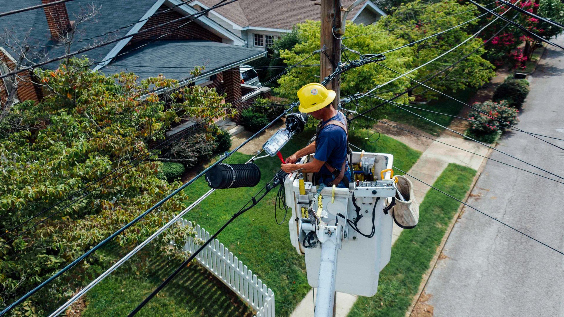 Utility worker in a bucket lift trimming tree branches near power lines along a residential street