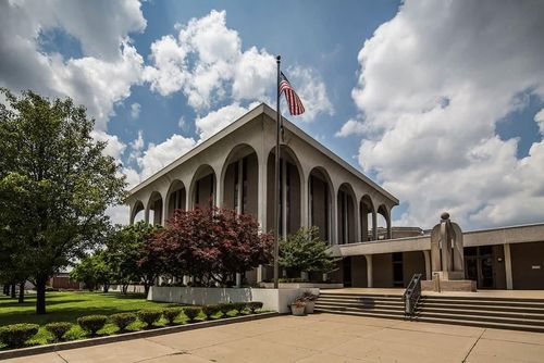 Modern civic building with arched façade, American flag, trees, and cloudy sky