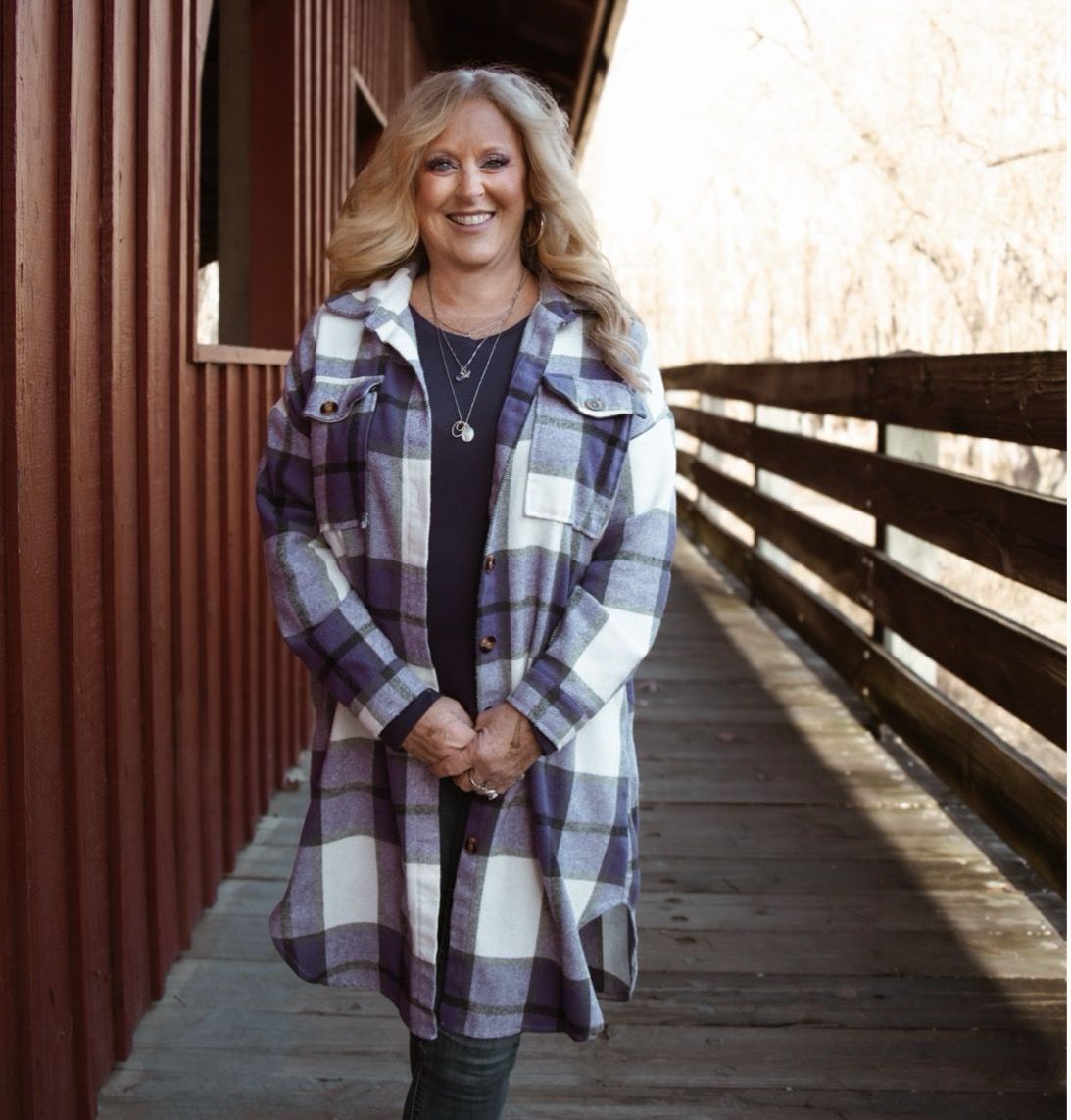 Woman in purple plaid jacket stands on a wooden bridge, smiling.