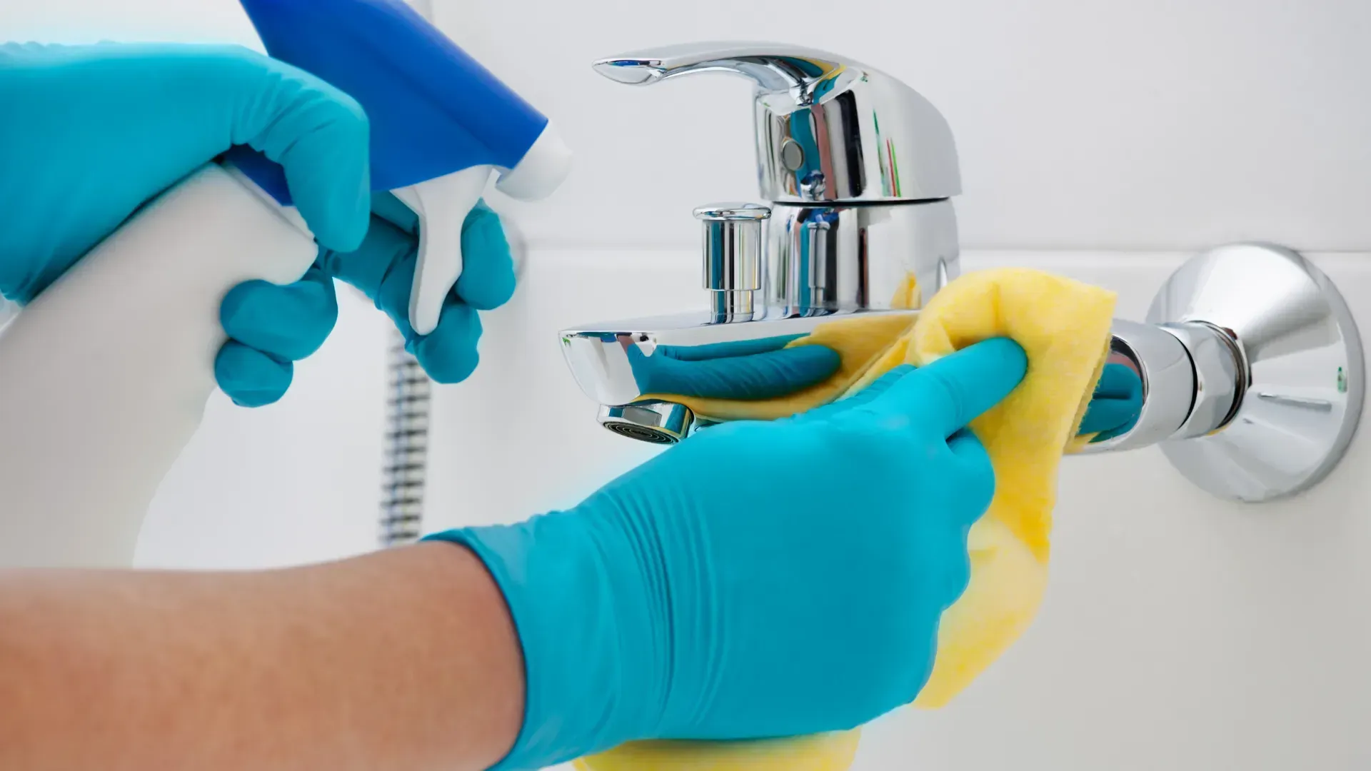 Person wearing blue gloves spraying and wiping a chrome faucet in a bathroom.