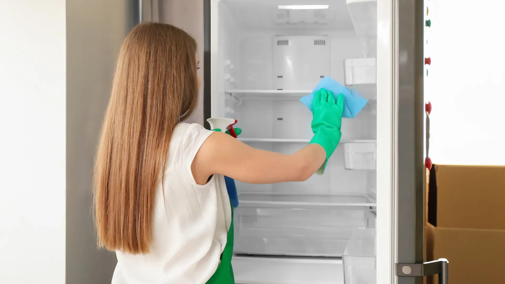 Woman wearing gloves cleans the inside of a refrigerator with a spray bottle and cloth.