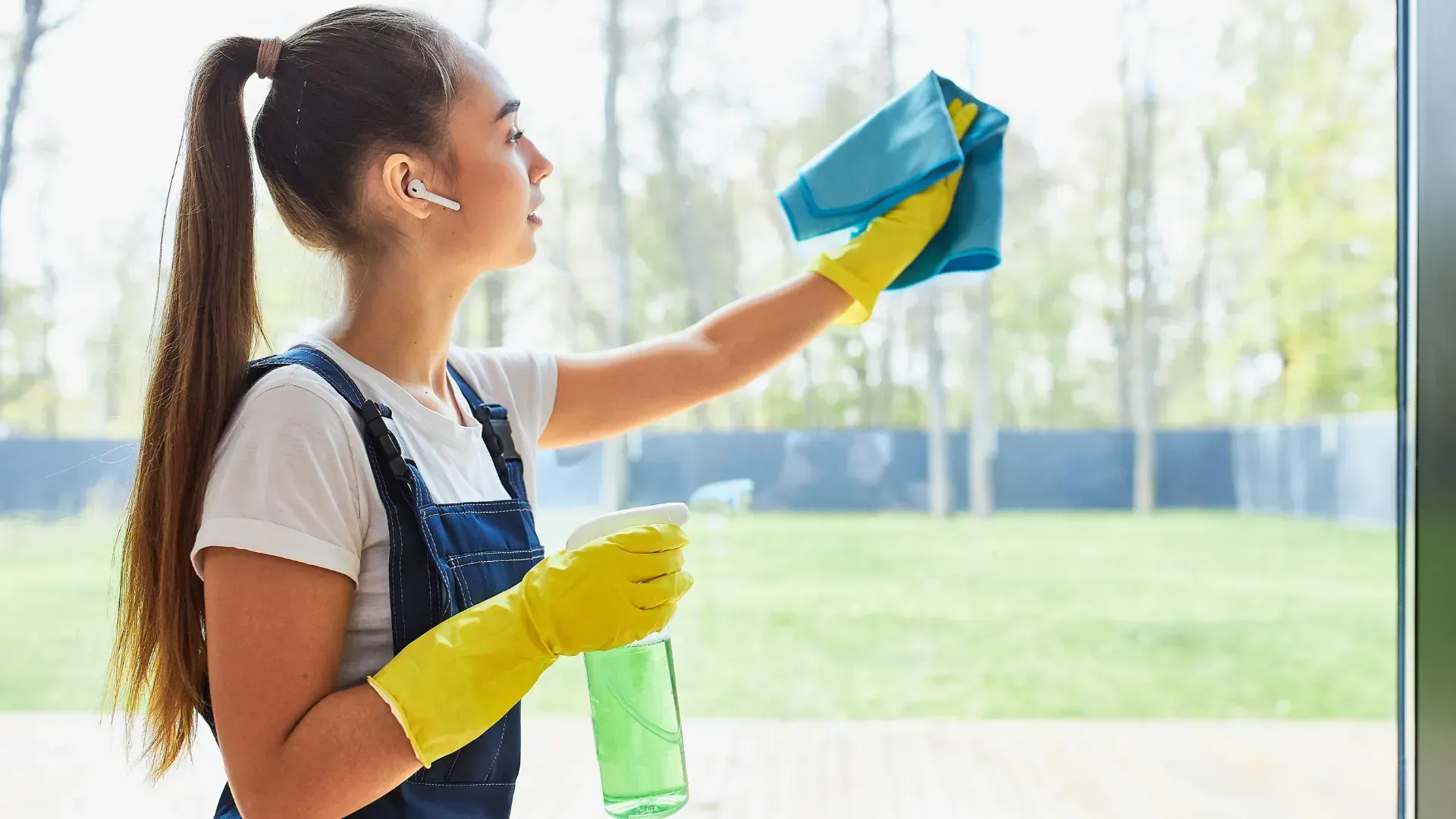 Woman wearing yellow gloves cleaning window with spray bottle and cloth; outdoors.