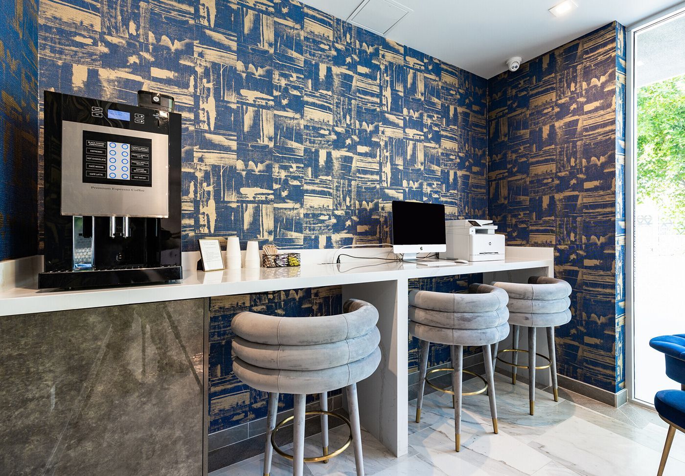 Coffee station with espresso machine, computer, stools, and patterned blue and gold wallpaper.