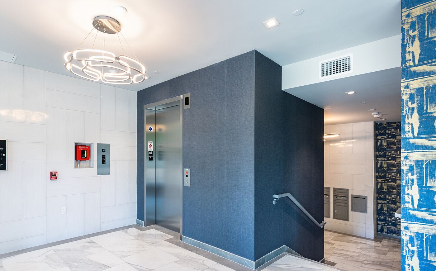 Elevator in modern building lobby with blue textured walls, silver fixtures, and decorative lighting.