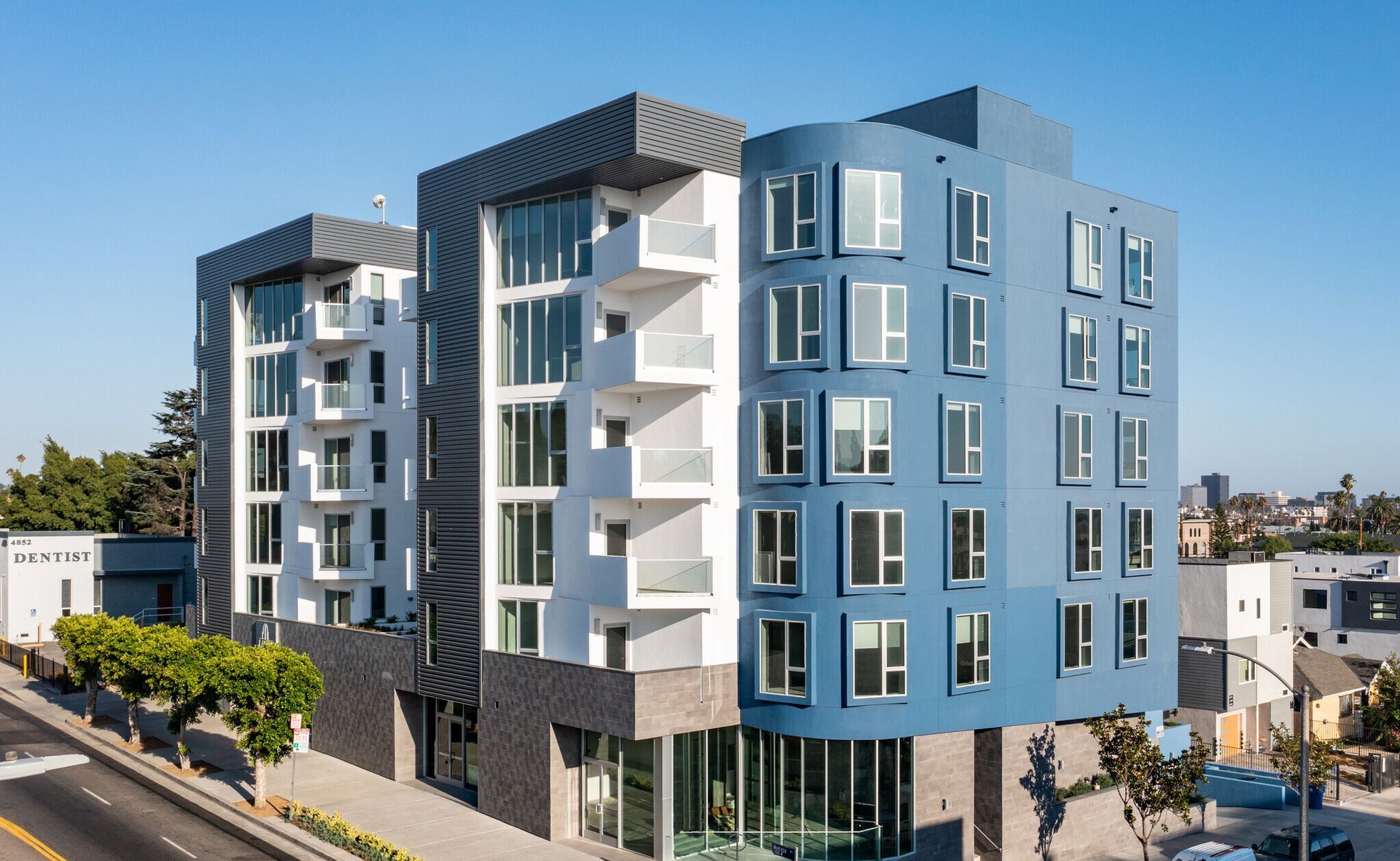 Modern multi-story apartment building, blue and white facade, curved corner, balconies, sunny day.