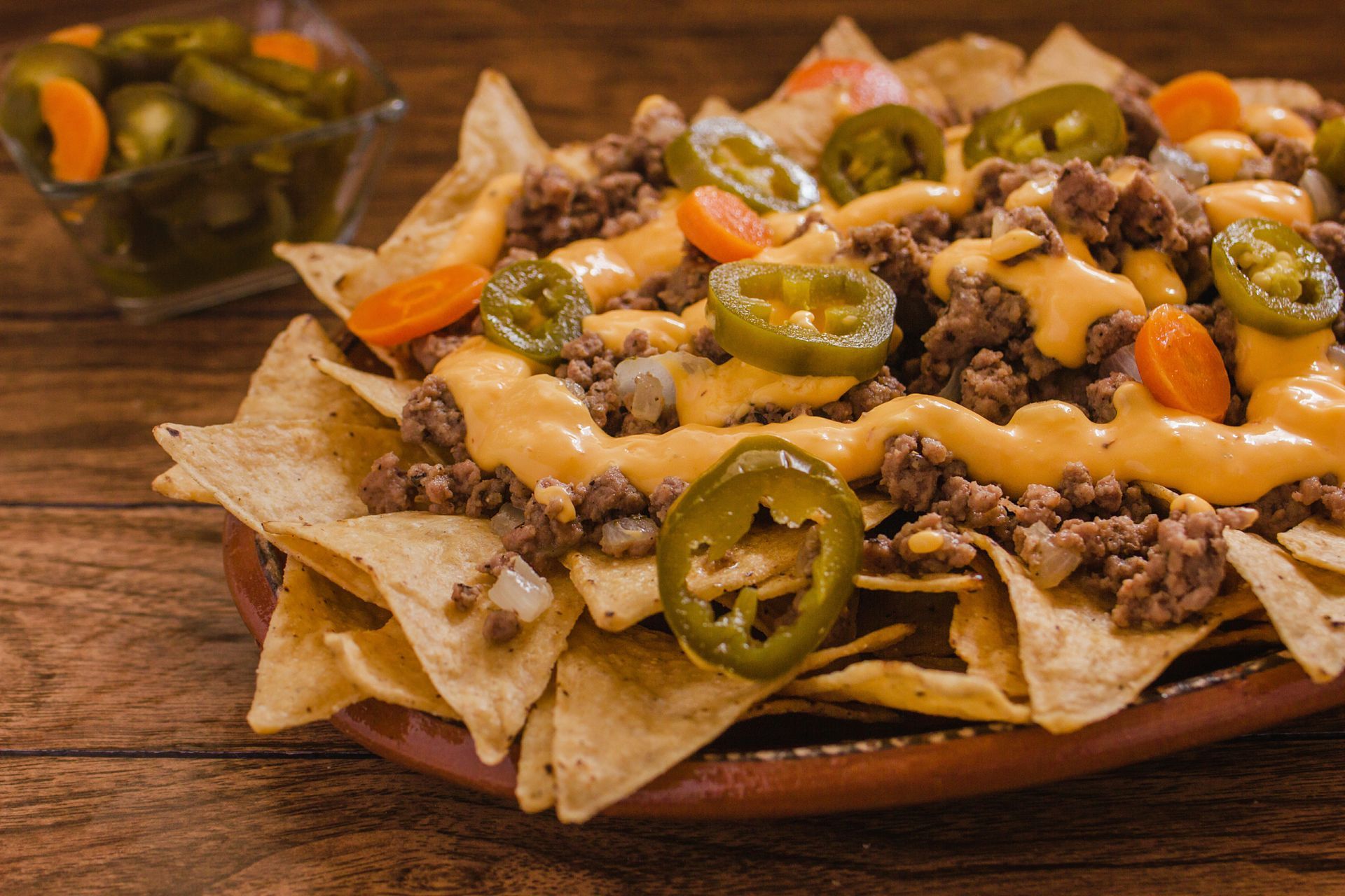 A plate of nachos with meat , cheese , jalapenos and carrots on a wooden table.