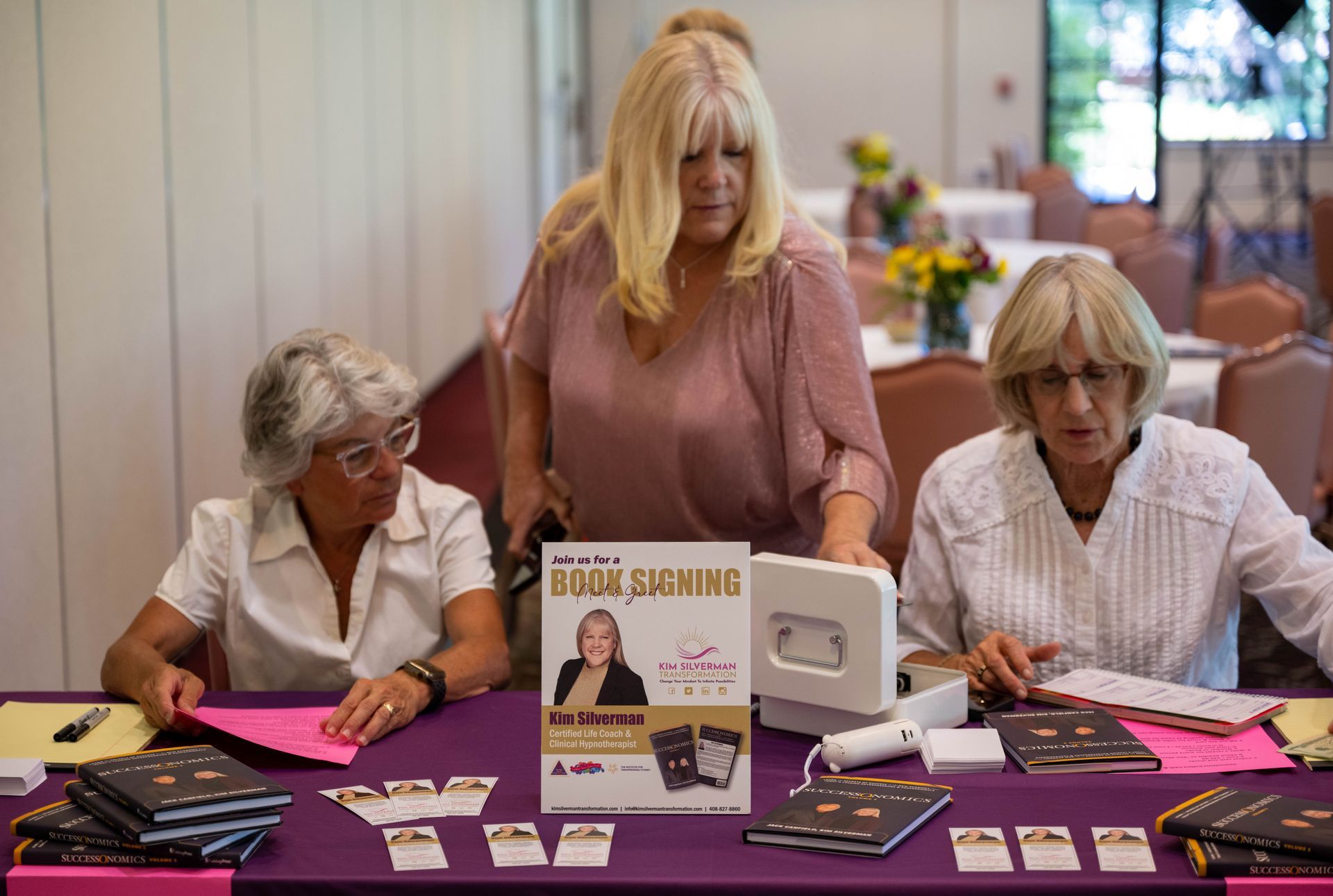 Three women are sitting and standing at a table with a sign that says book signing.