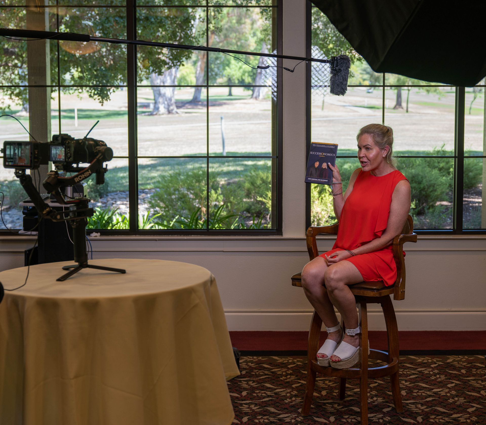 A woman in a red dress is sitting on a stool in front of a camera.