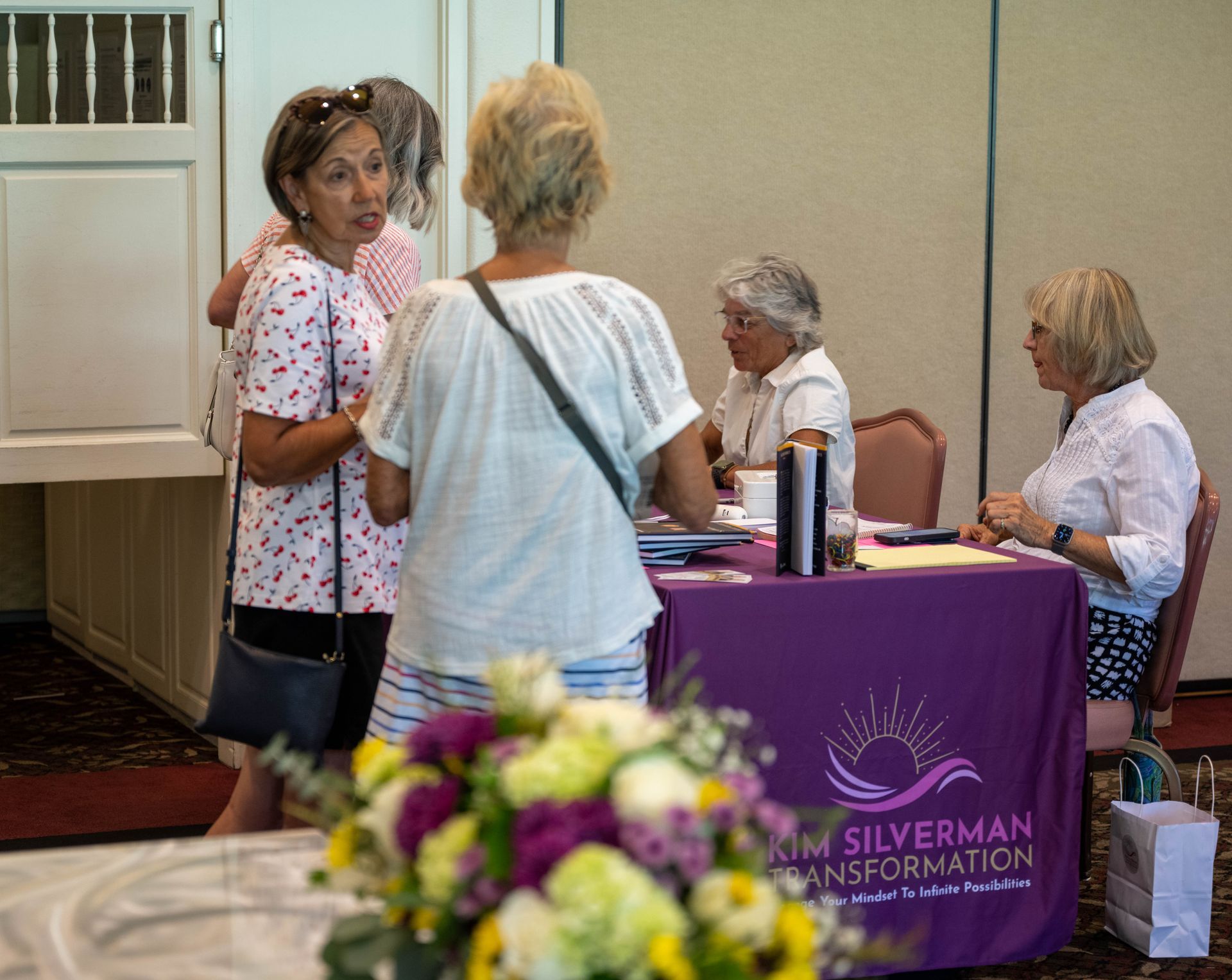 A group of women are standing around a table with a purple table cloth that says kim silverman transformation.