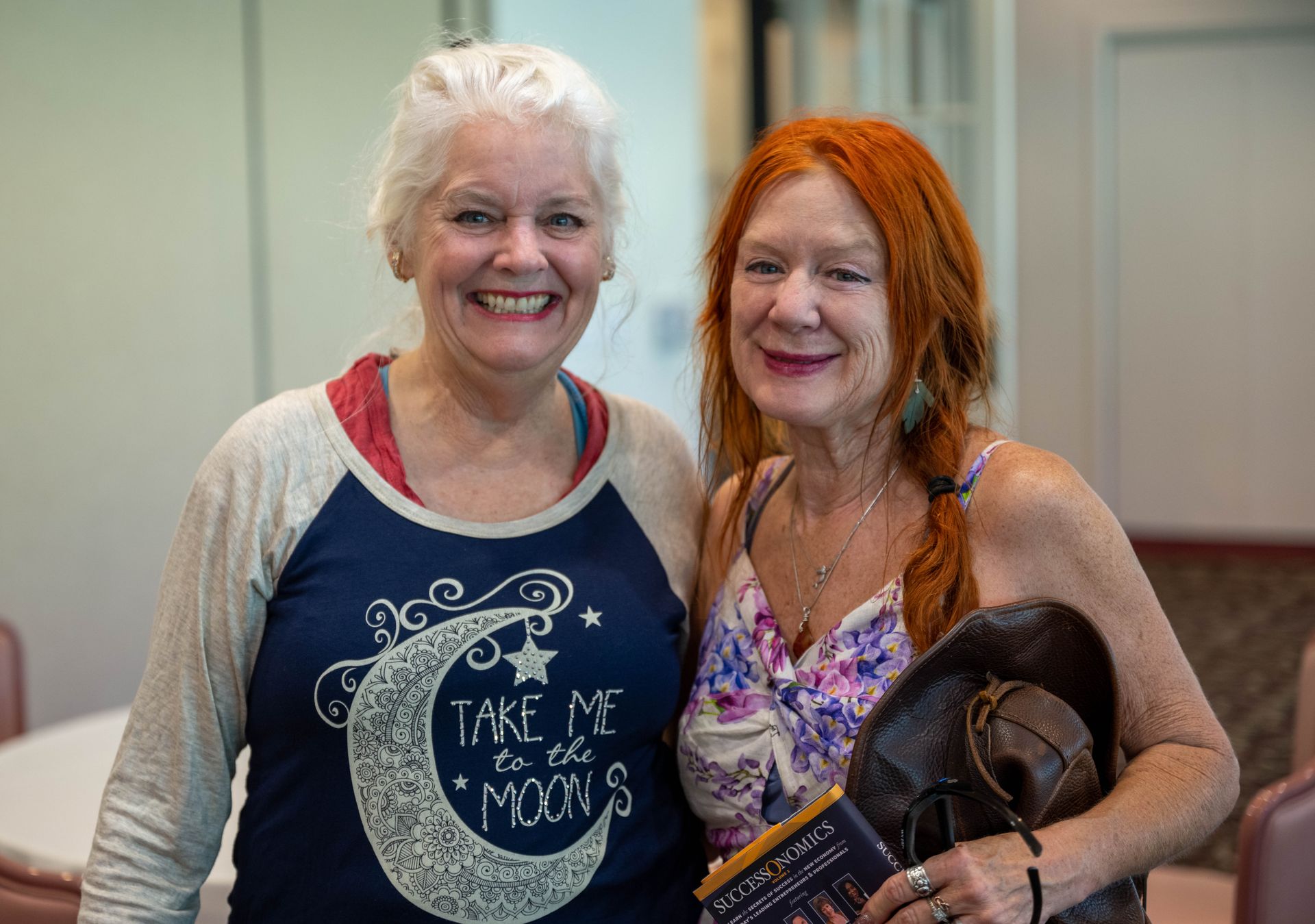 Two women are posing for a picture together and one of them is wearing a shirt that says `` take me to the moon ''.